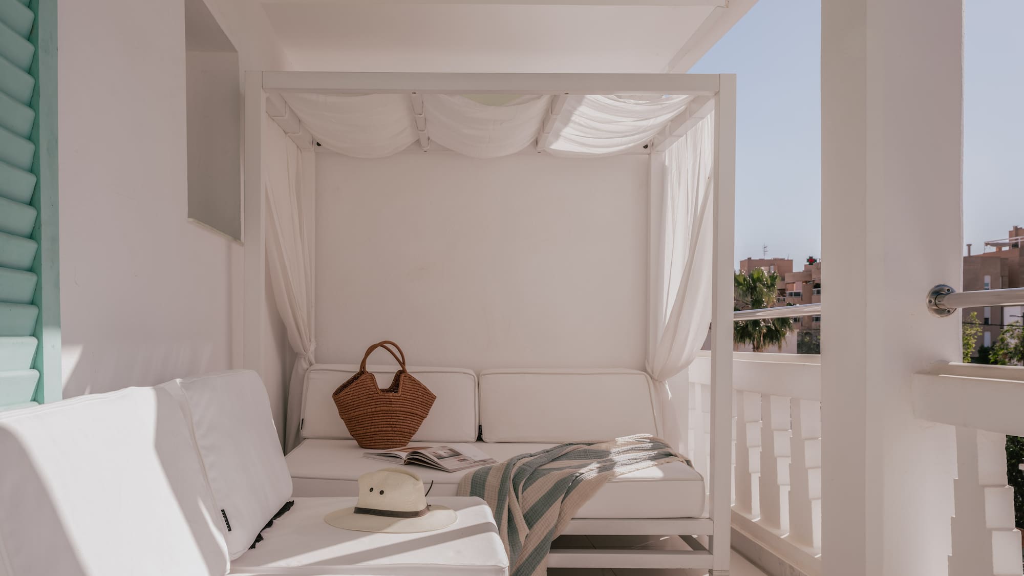 White hotel balcony with canopy daybed, lounge sofa, straw hat, and bag.