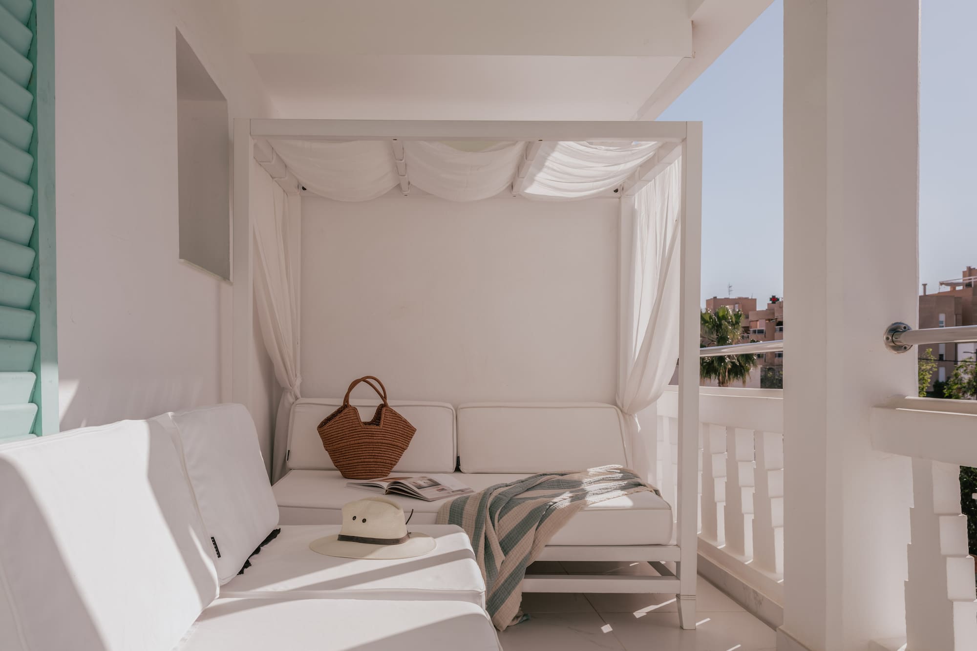 Sunny white balcony lounge with canopy daybed, straw hat, and blanket.