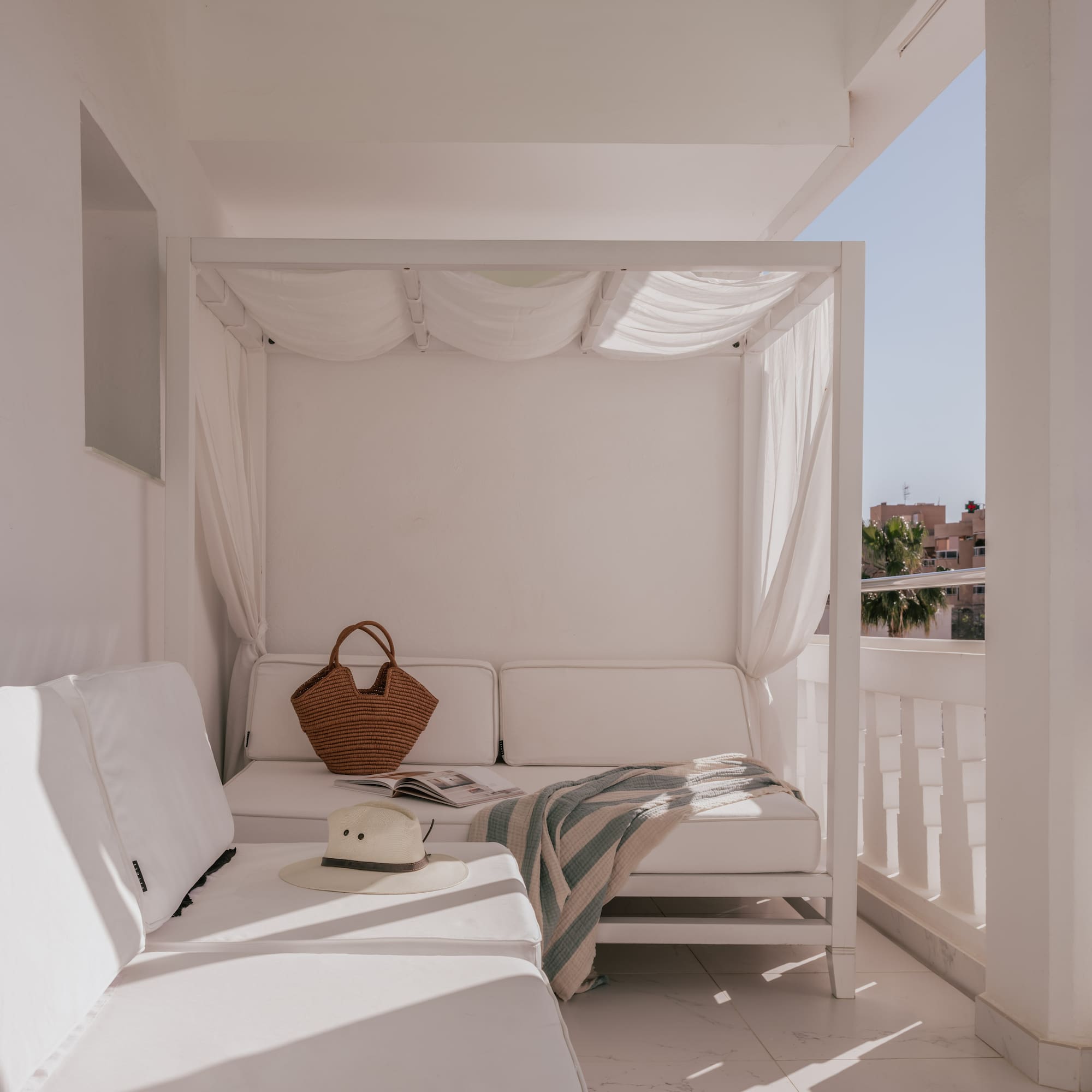 Sunny white balcony lounge with canopy daybed, straw hat, and blanket.