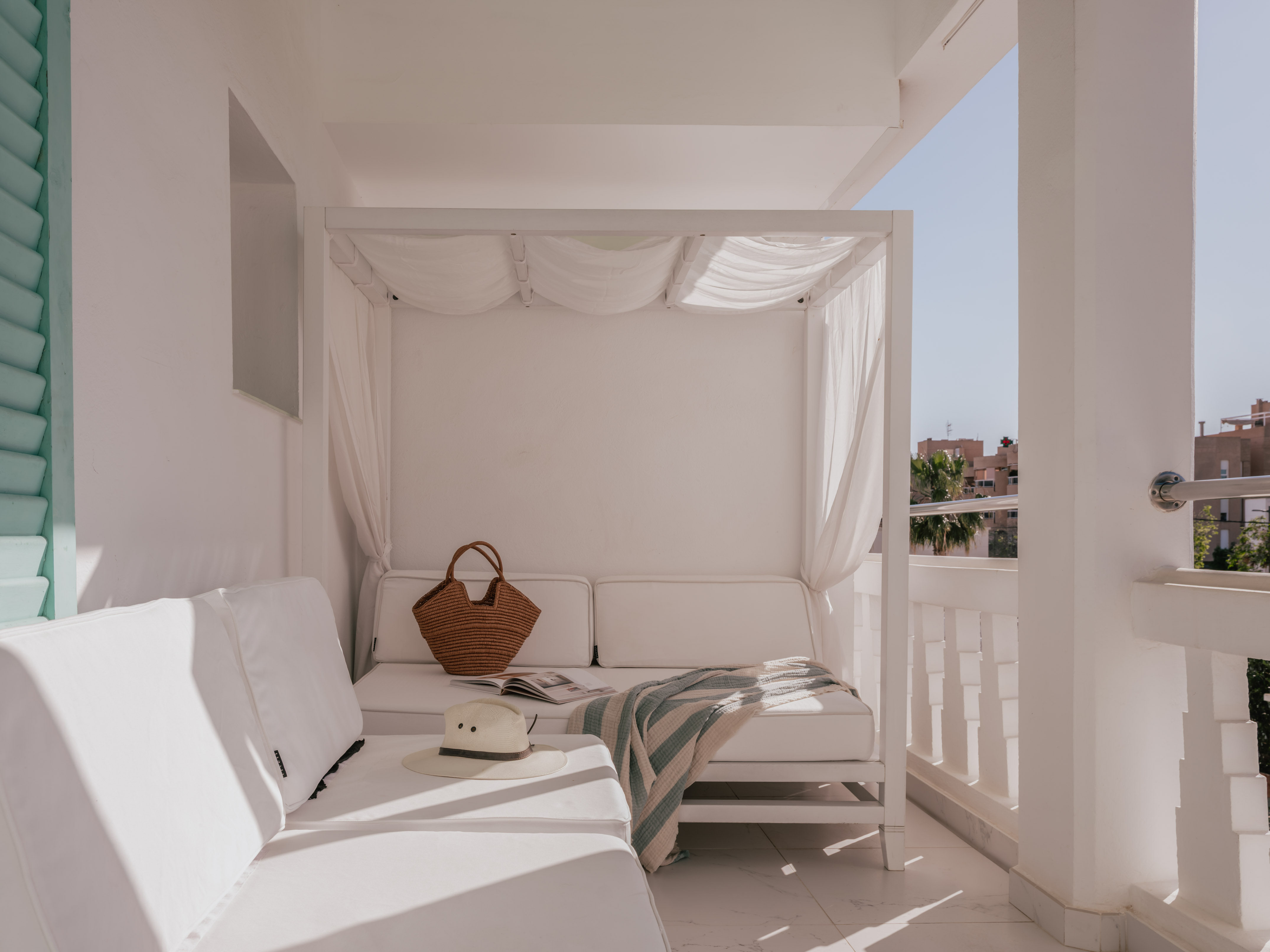 Sunny white balcony lounge with canopy daybed, straw hat, and blanket.