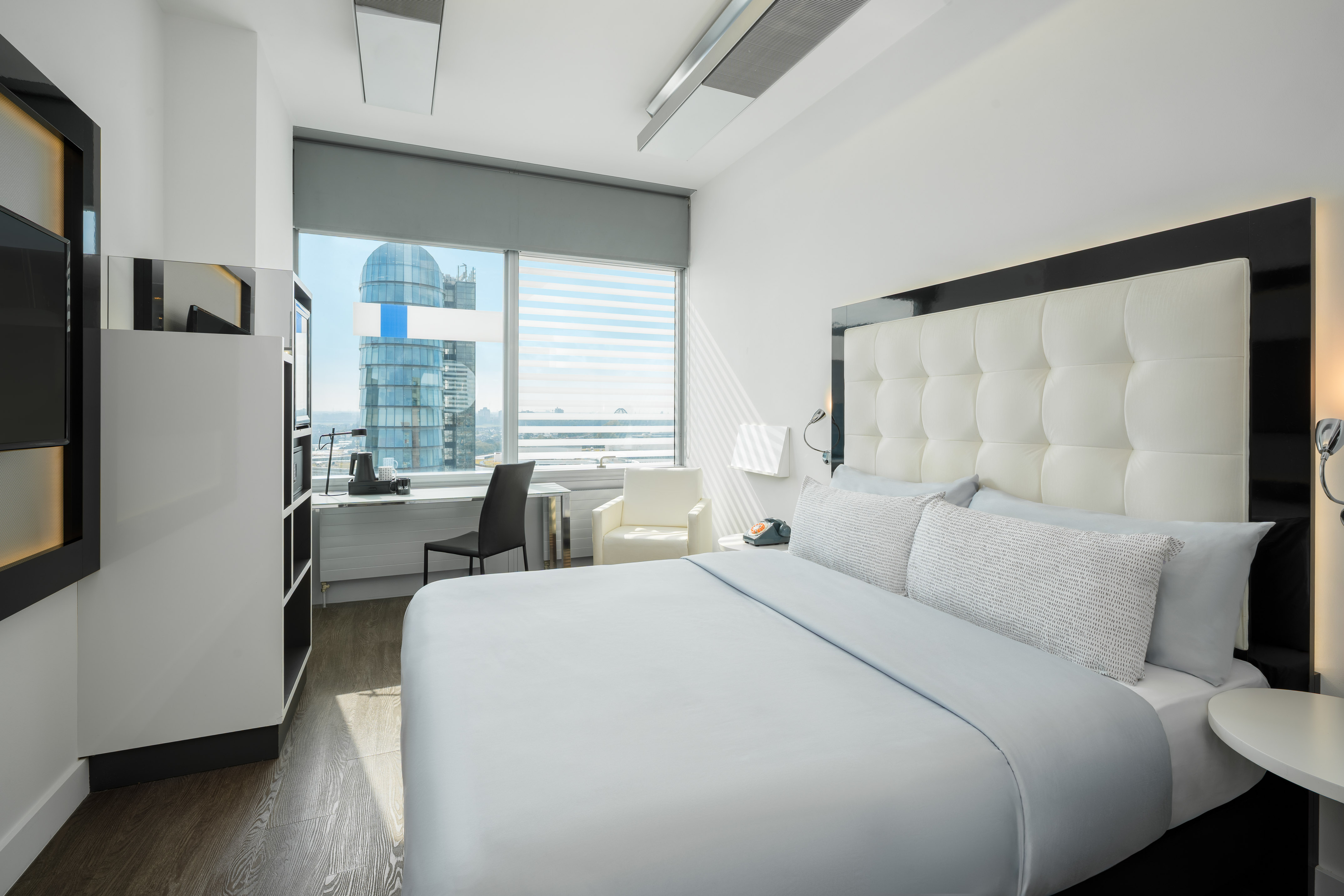 Modern hotel room with white bed, workstation, and cityscape view.