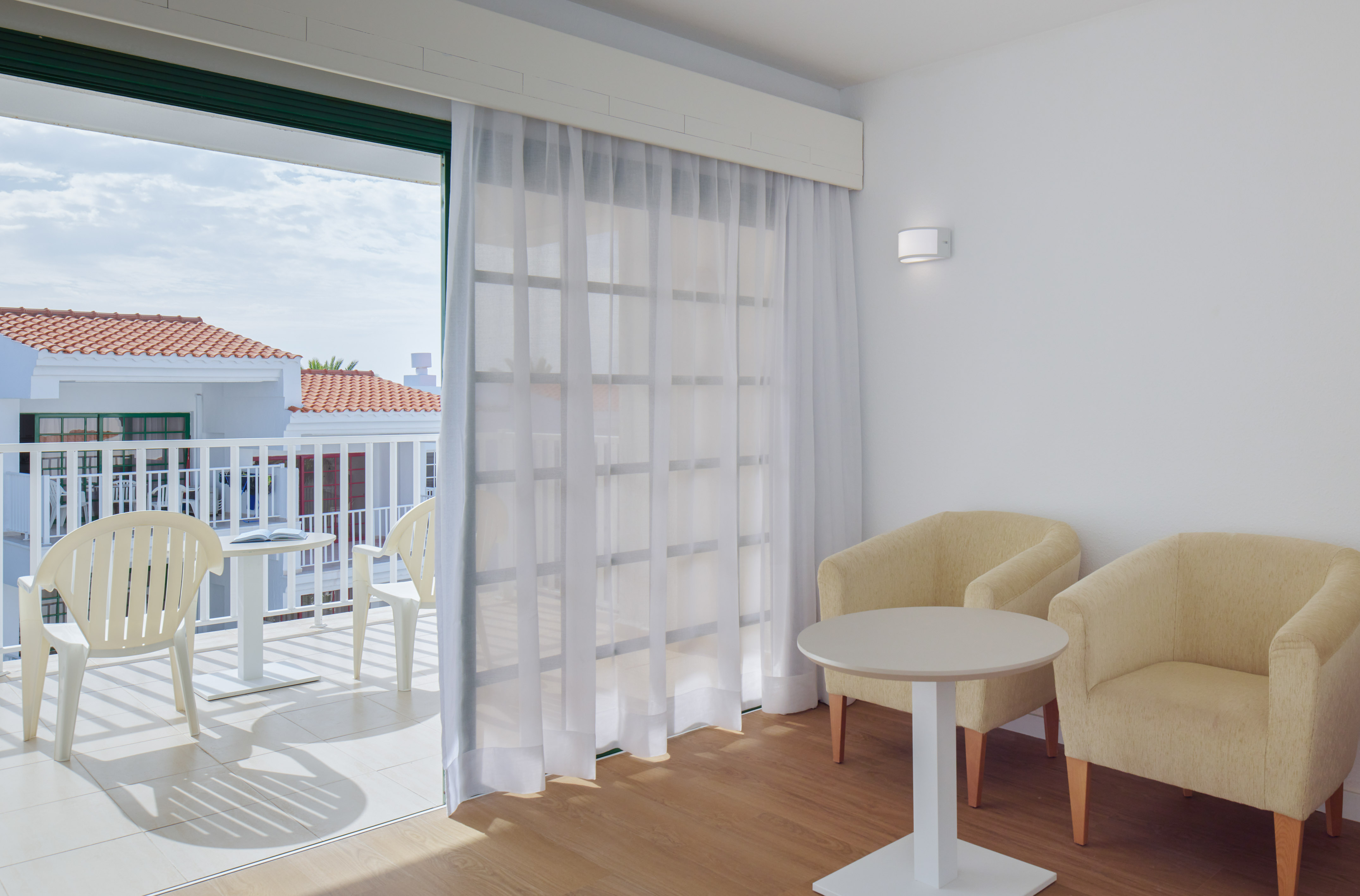 Bright hotel room with balcony access, armchairs, and view of tiled roofs.