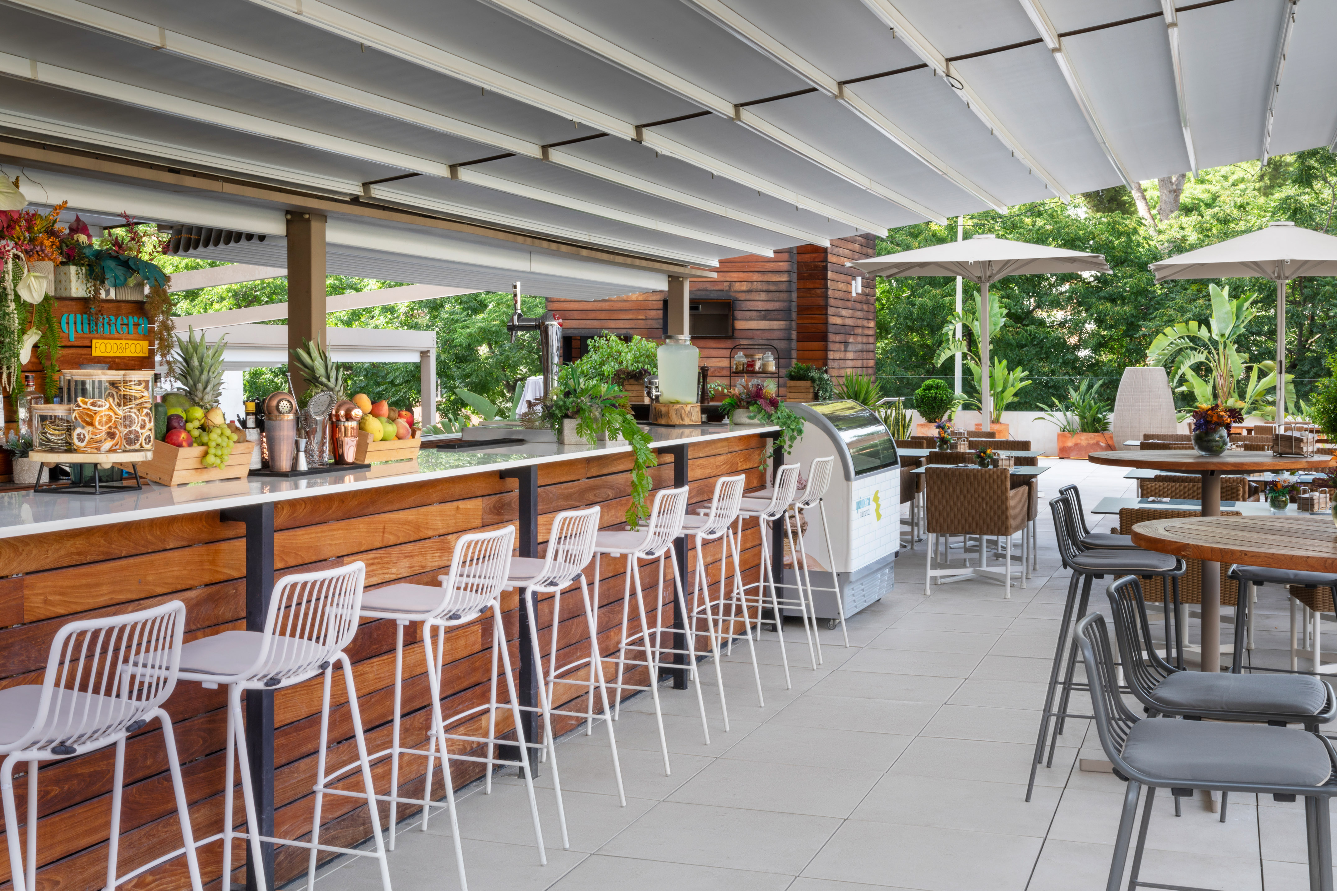 Outdoor hotel bar with fresh fruit, white bar stools, and shaded patio seating.