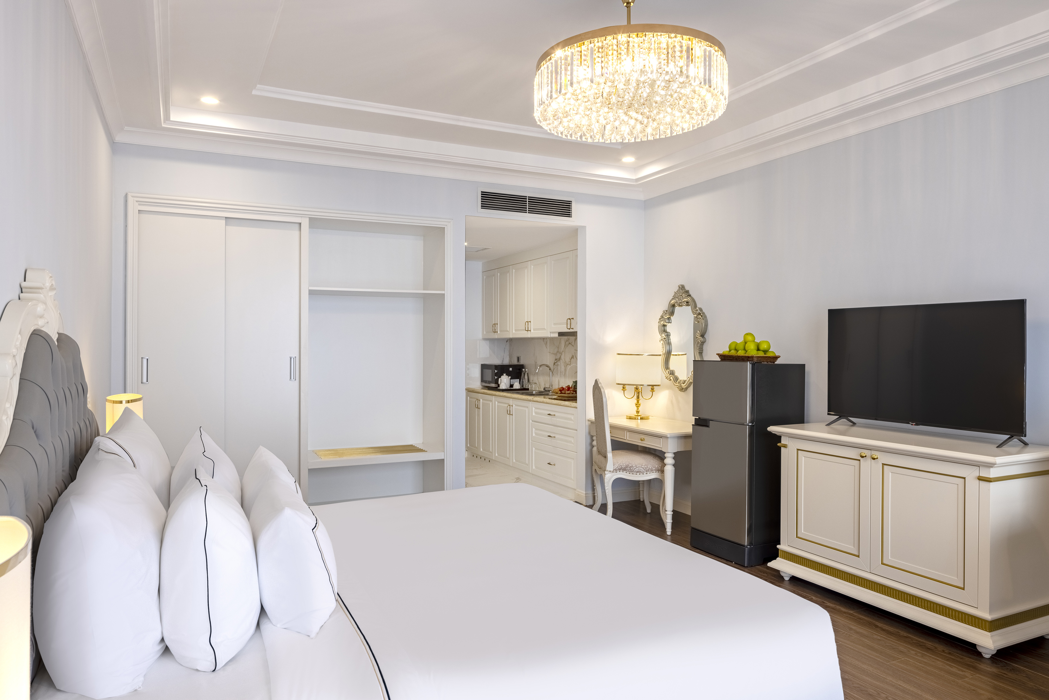 Elegant white hotel room with bed, TV, desk, and modern kitchenette area.