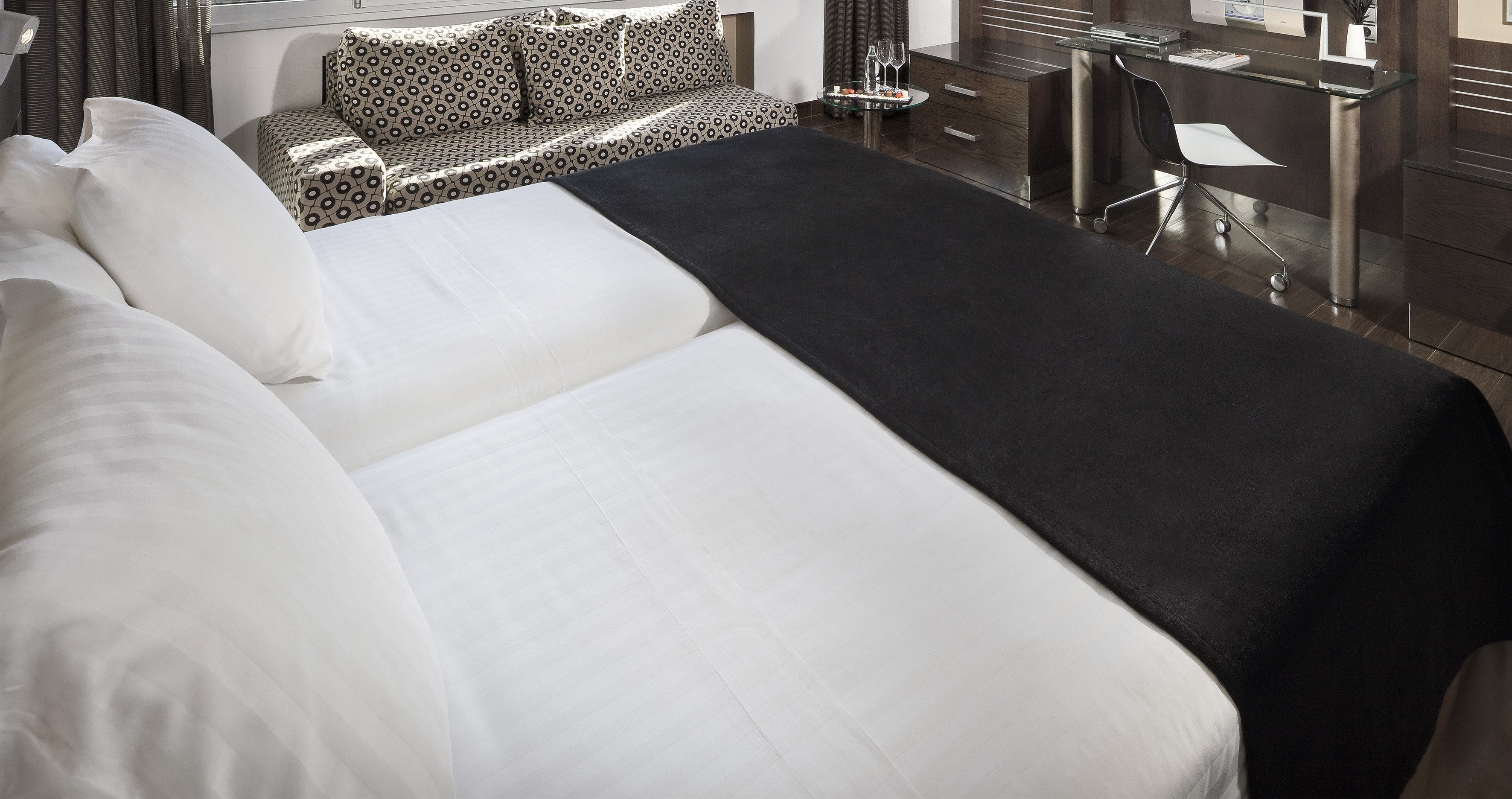 Modern hotel room with twin beds, white linens, black throw, sofa and desk.
