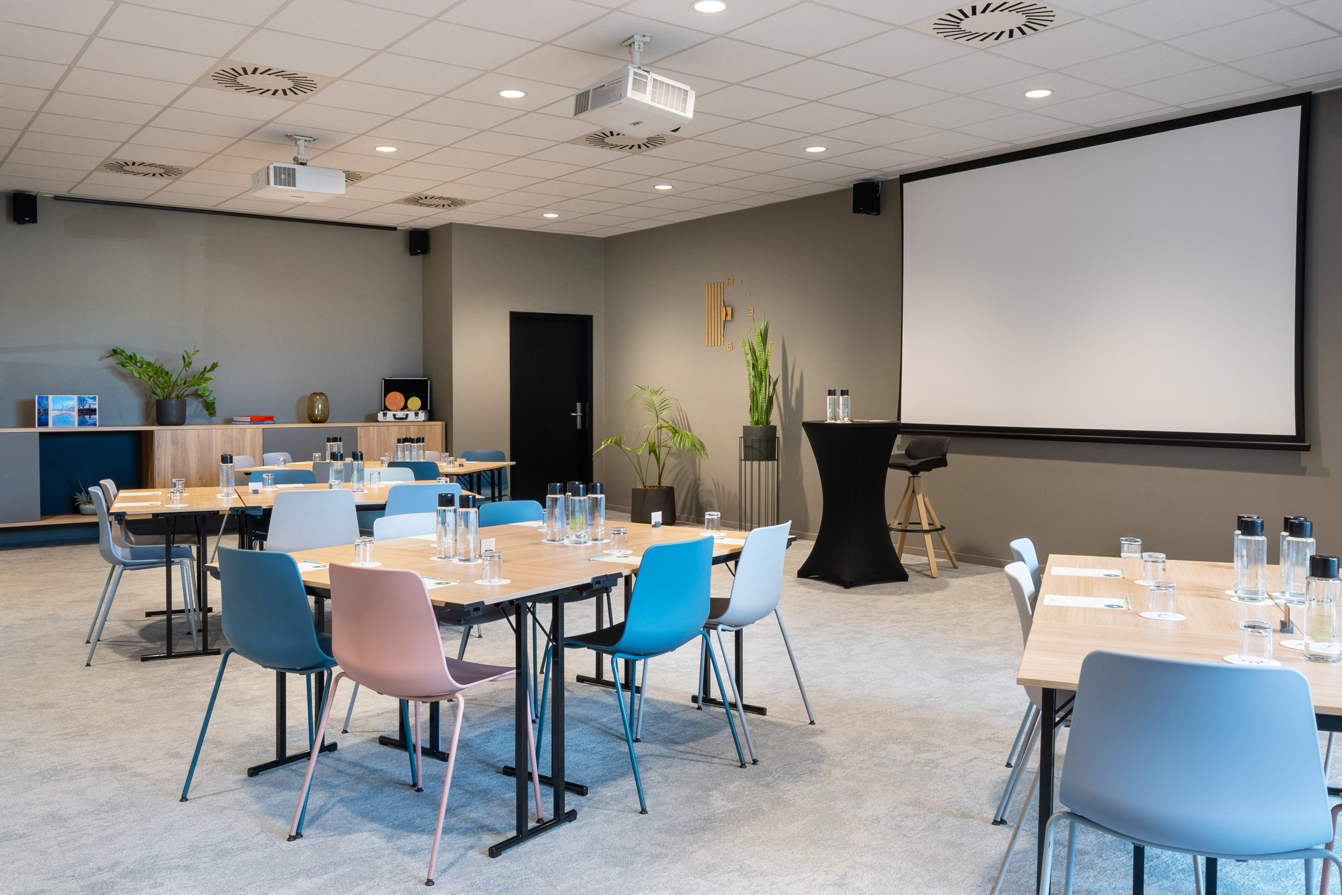 Modern meeting room with projector screen, wooden tables, and colorful chairs.