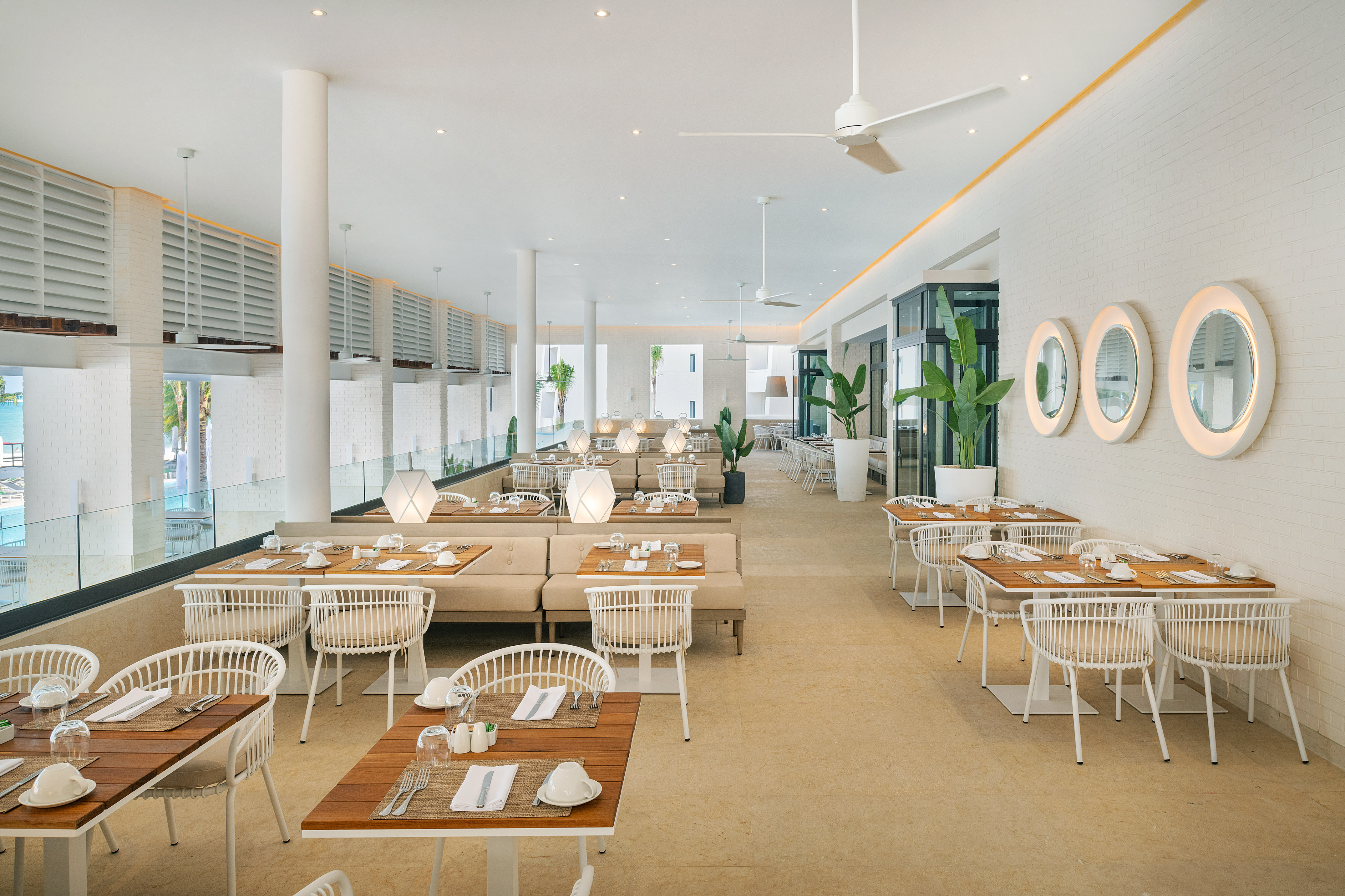 Bright modern restaurant with white seating, wooden tables, and tropical plants.
