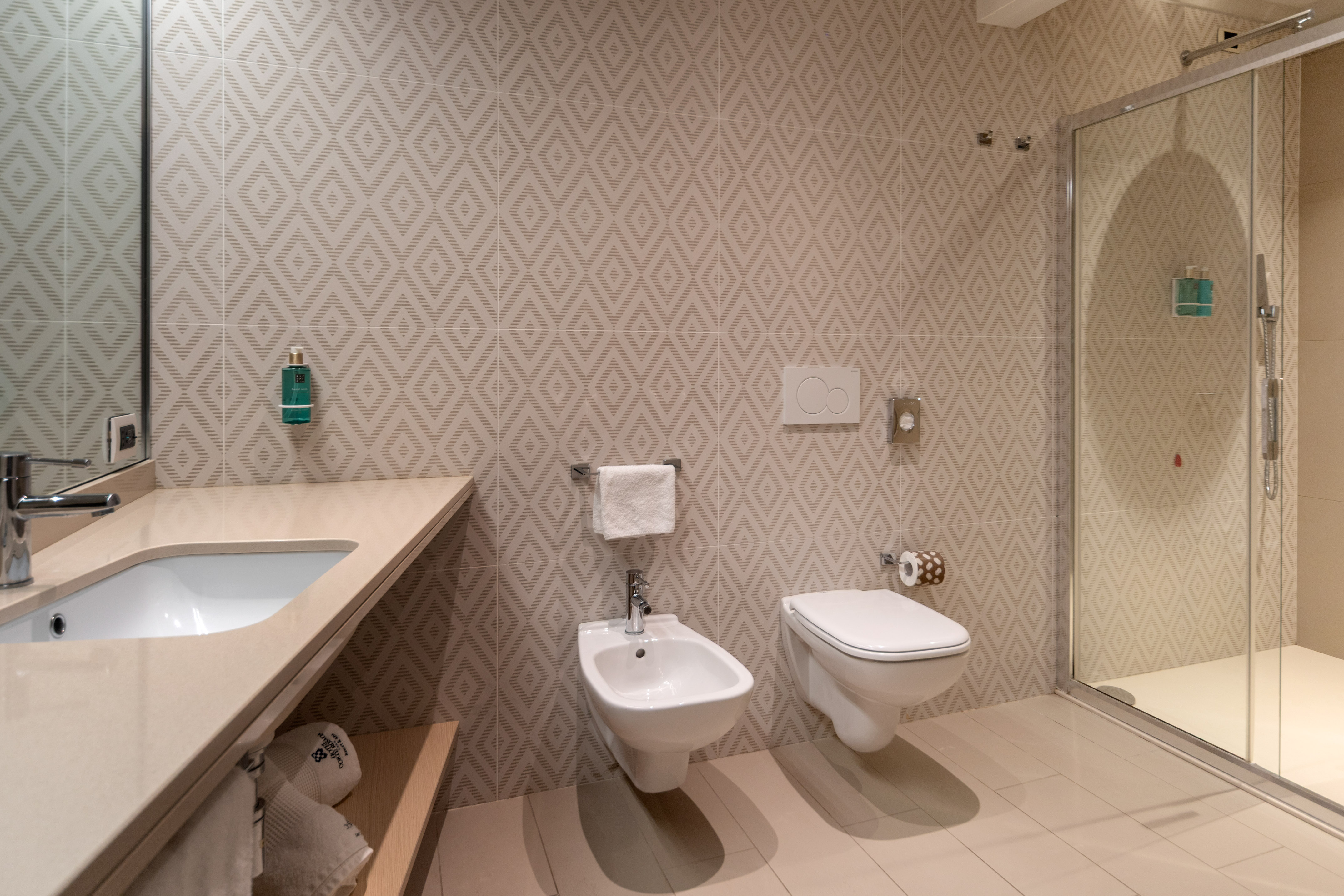 Modern hotel bathroom: patterned tiles, vanity sink, toilet, bidet, glass shower.