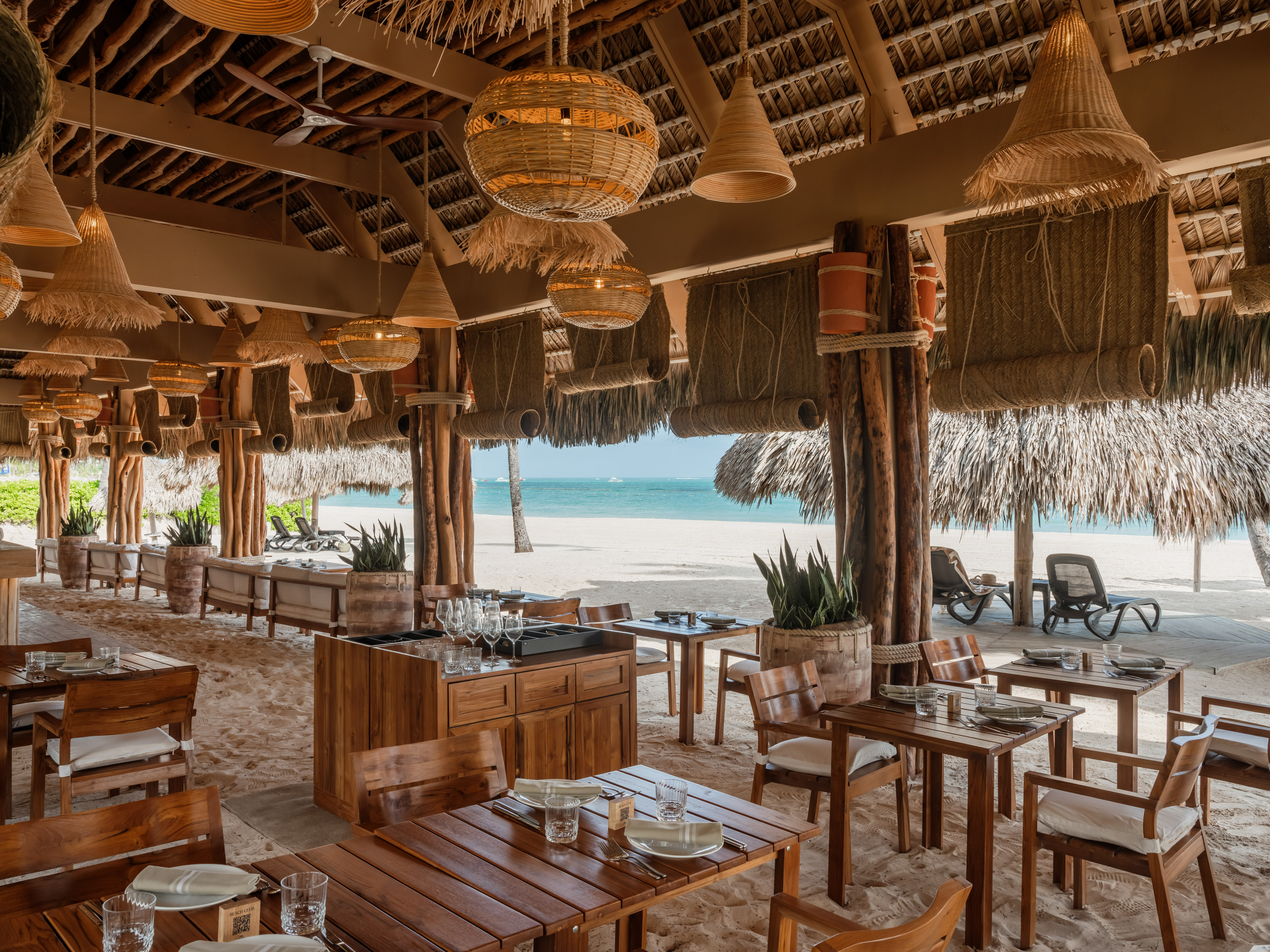 Beachfront restaurant, wooden tables, woven lamps, thatch roof, and ocean view.