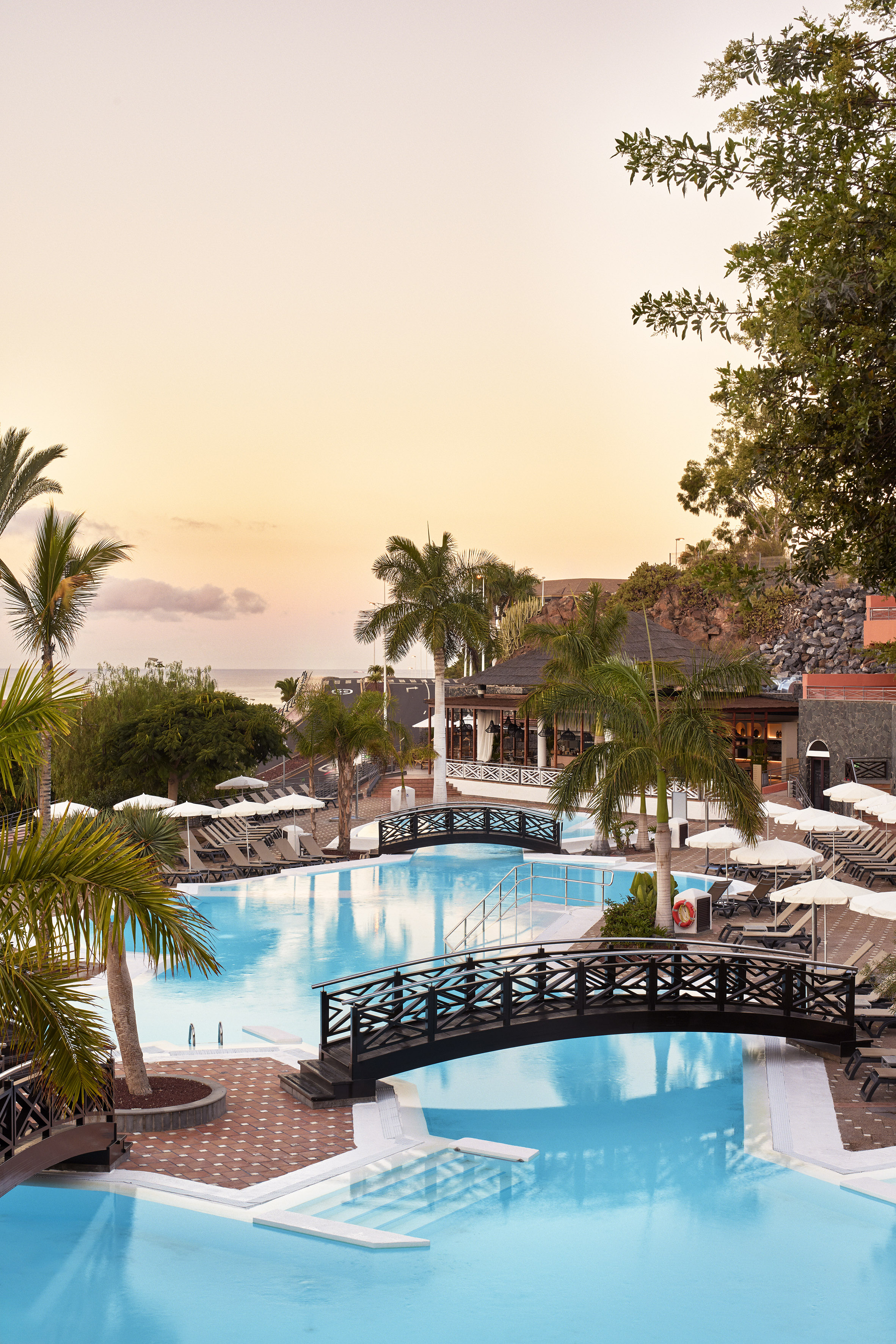 Sunset over tropical resort pool with bridges, palm trees, ocean view.