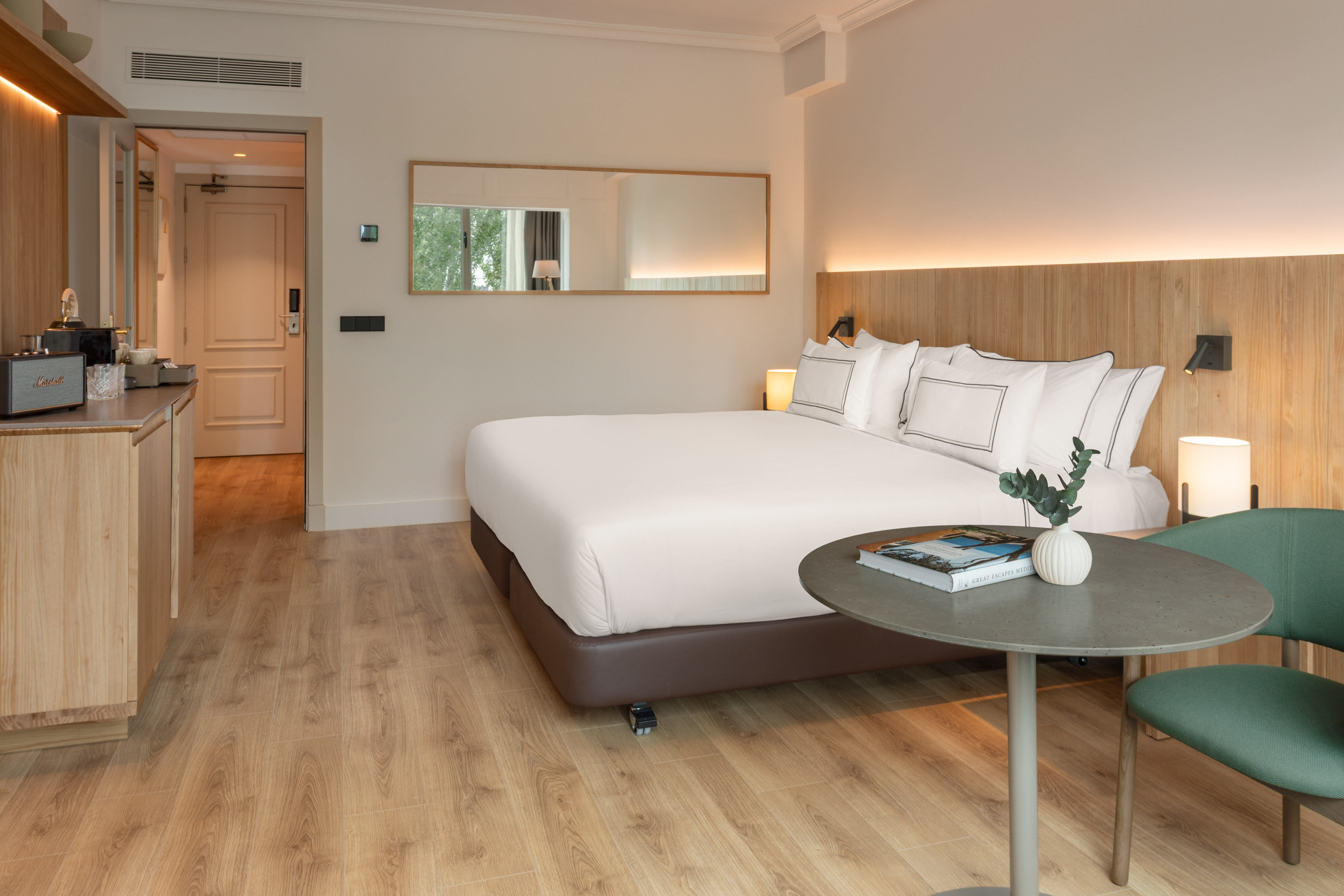 Modern hotel room: king bed, wood headboard, light floor, and green chair.