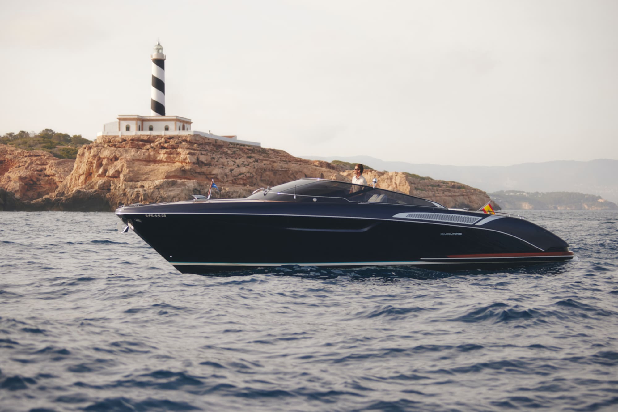 Dark blue luxury yacht cruising past a striped lighthouse on a rocky island.