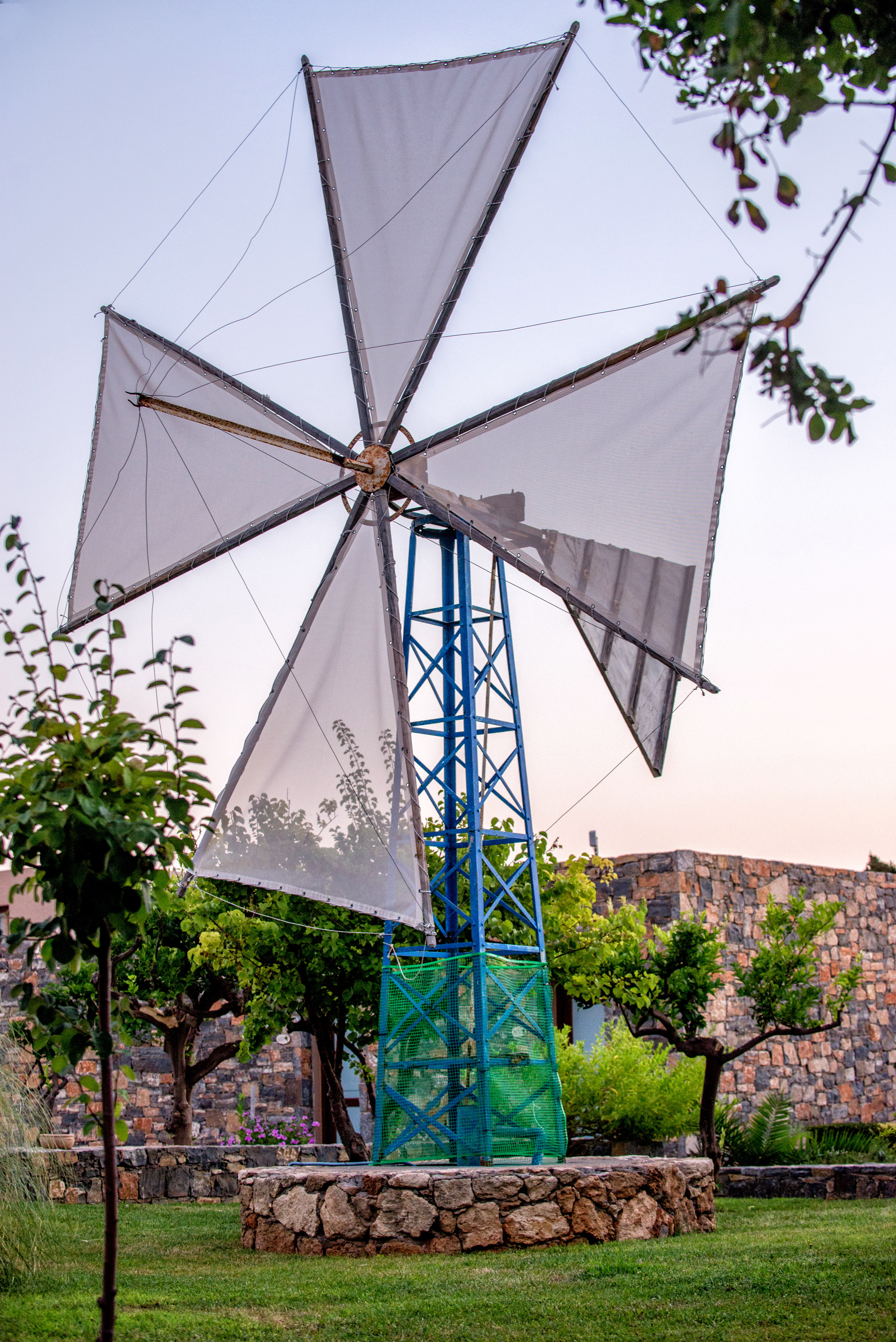 Cretan windmill with blue base and white sails in a green garden.