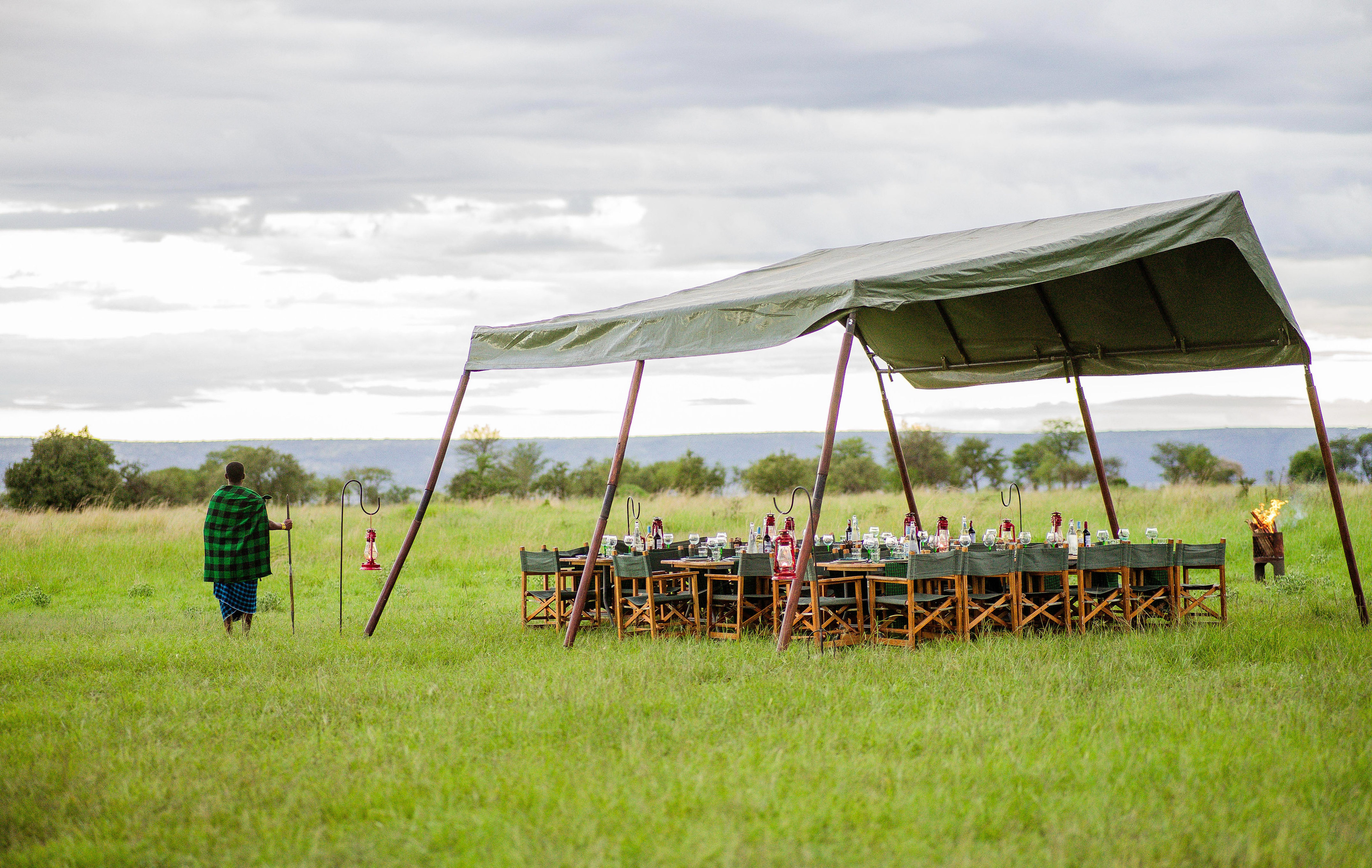 Outdoor safari dinner in savanna with canopy, campfire, and Maasai guide.