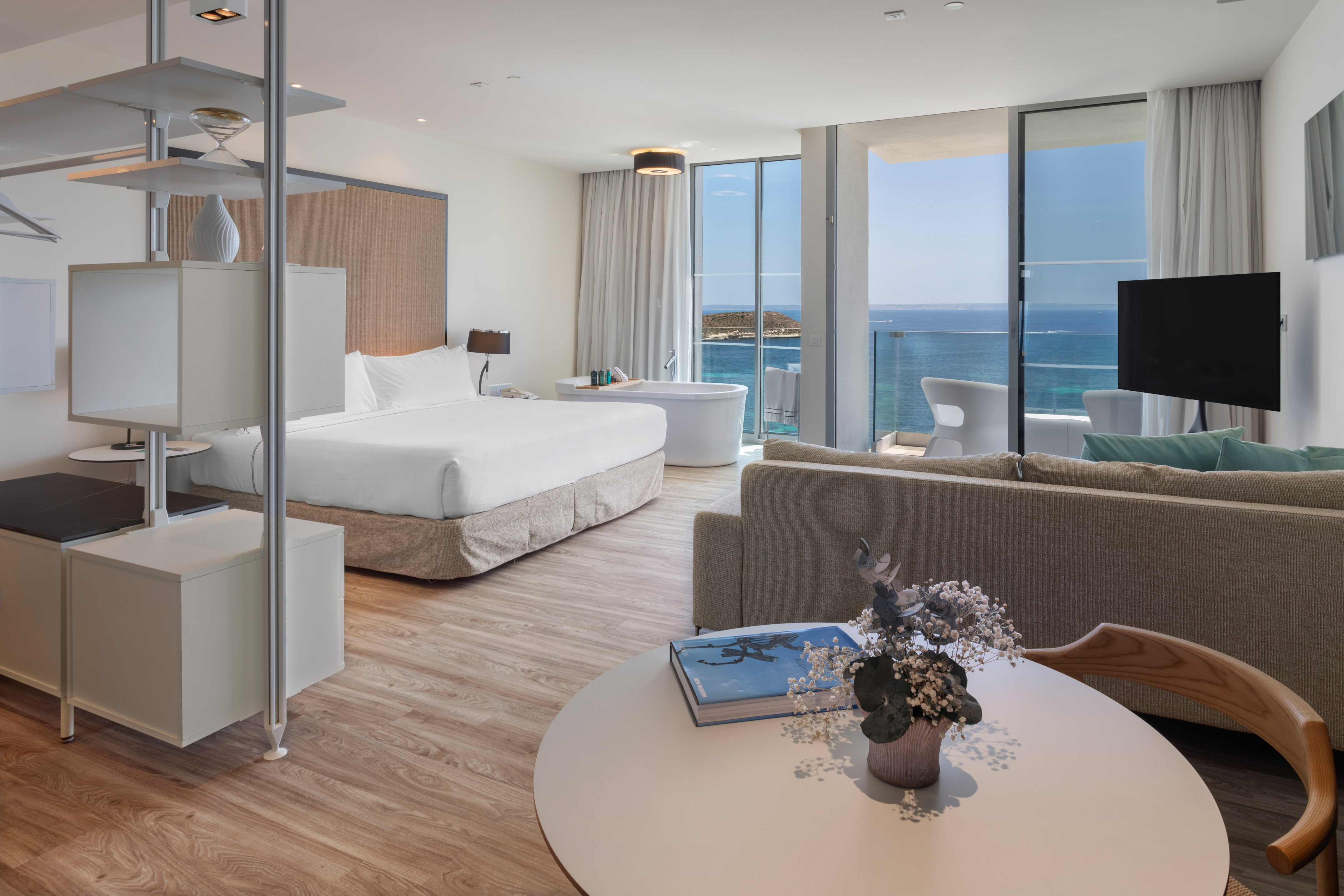 Modern hotel suite with ocean view balcony, bed, sofa, and freestanding tub.