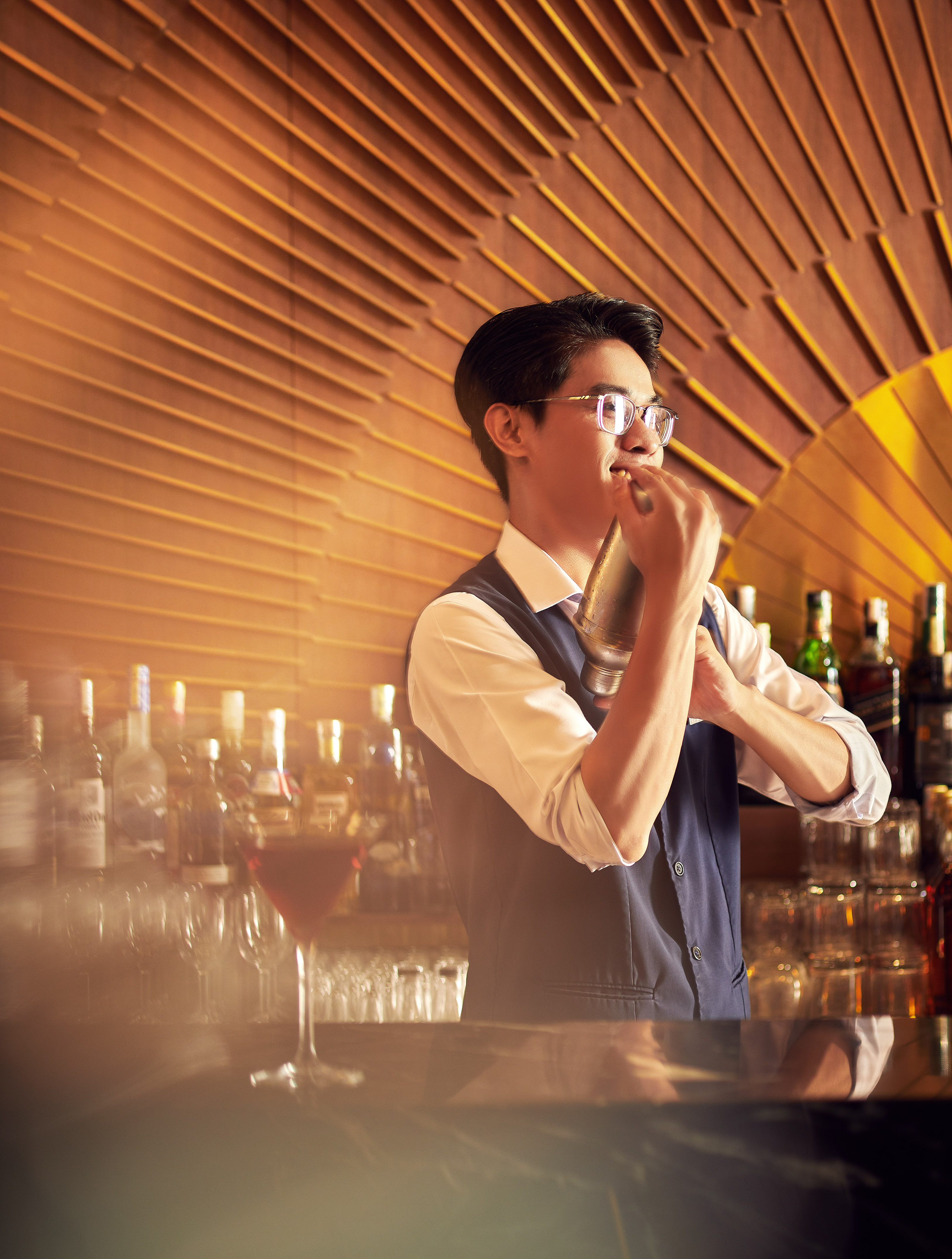 Bartender in glasses shaking a cocktail shaker behind a bar with bottles.