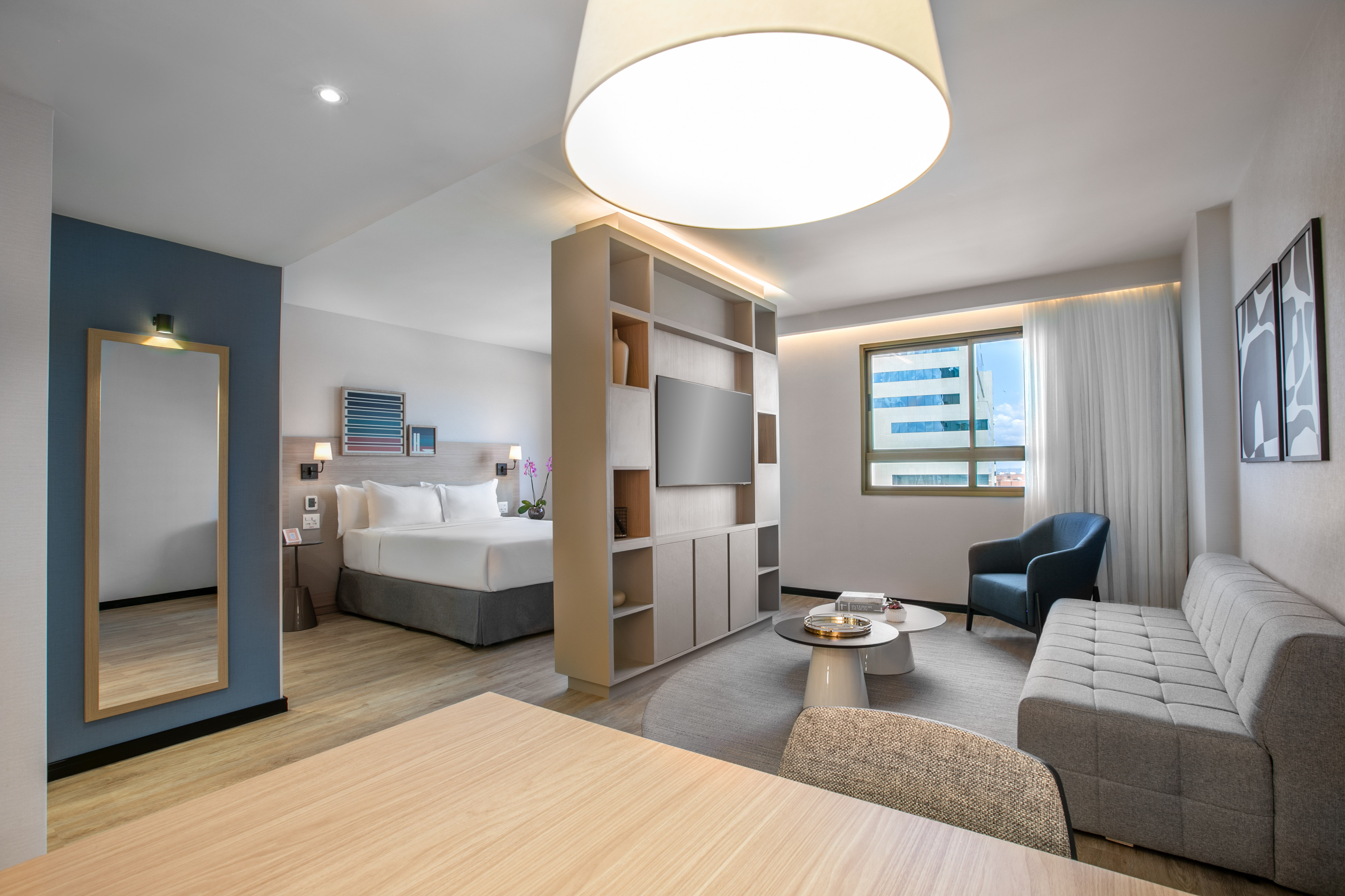Modern hotel studio suite with king bed, sofa, TV, and city view window.