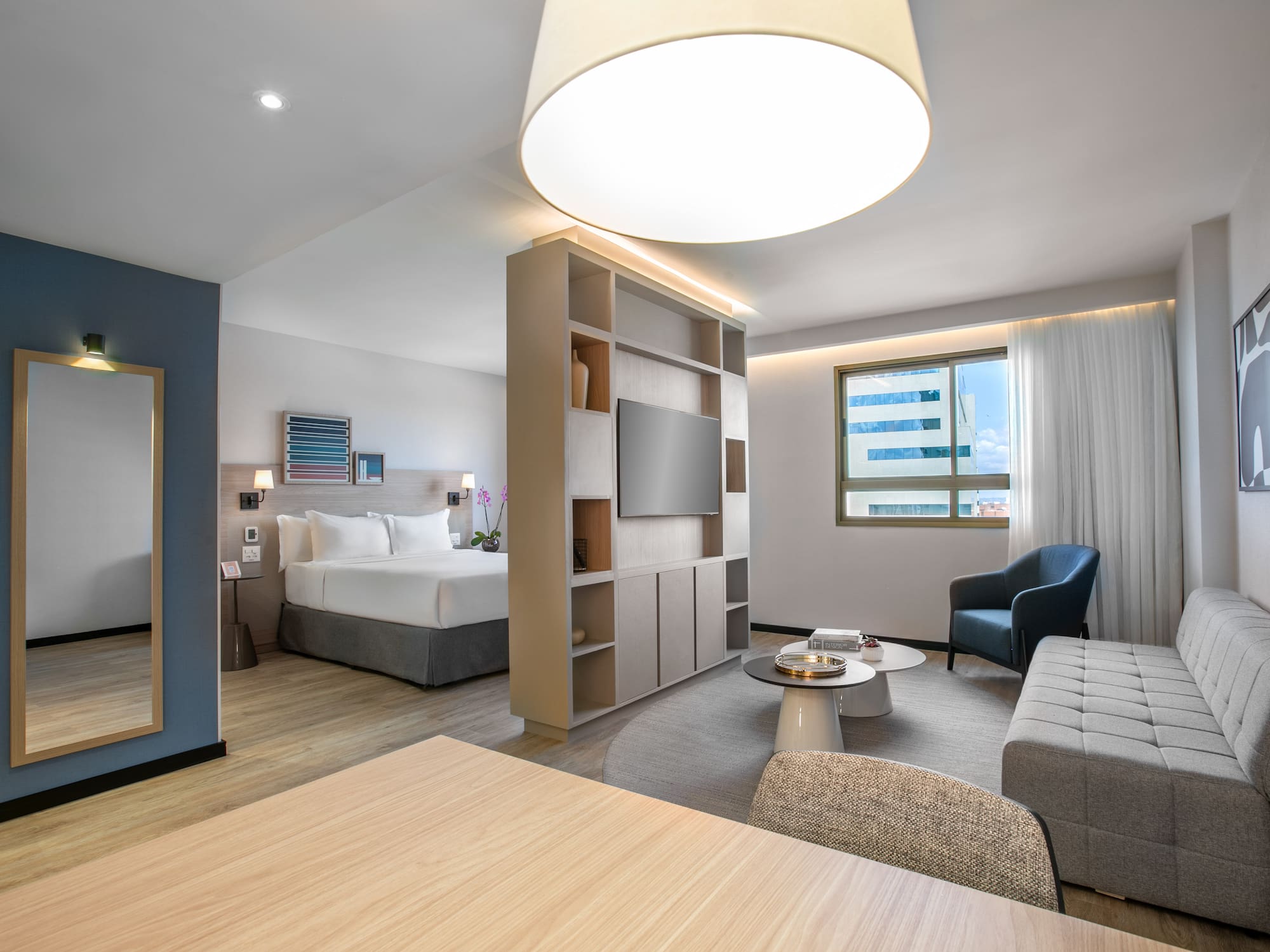 Modern hotel studio suite with king bed, sofa, TV, and city view window.