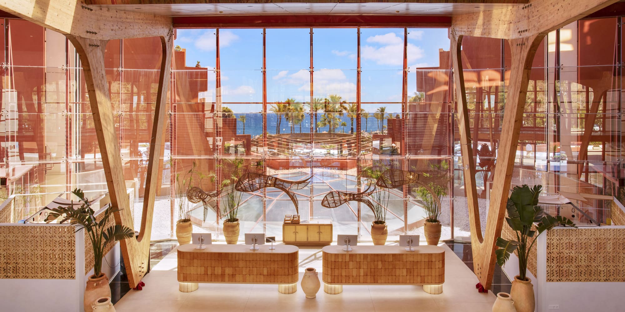 Modern hotel lobby with ocean view, wooden reception desks, and tropical plants.