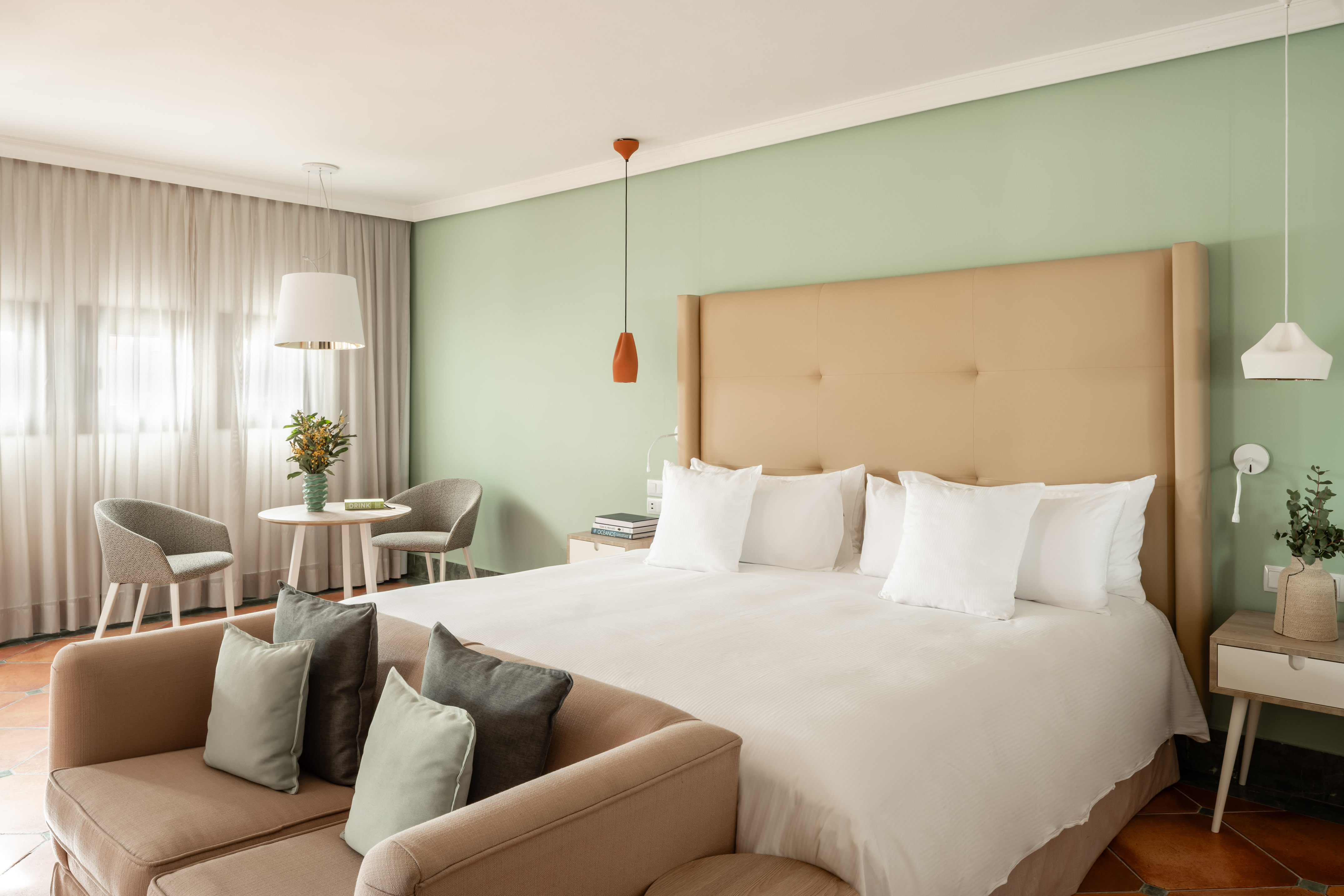 Hotel bedroom with large bed, beige headboard, light green walls, and sofa seating.
