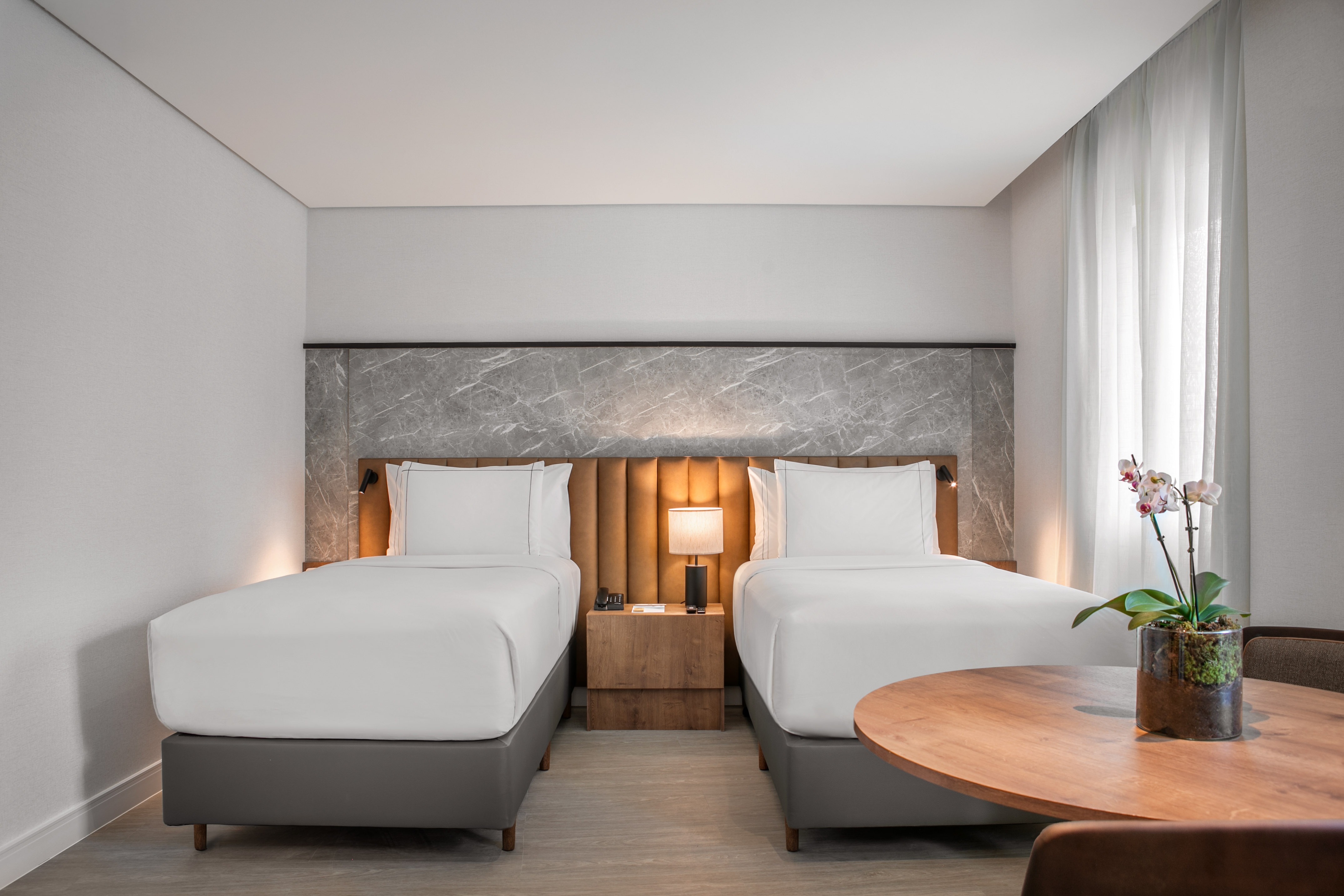 Modern hotel room with two twin beds, marble headboard, and wood table.