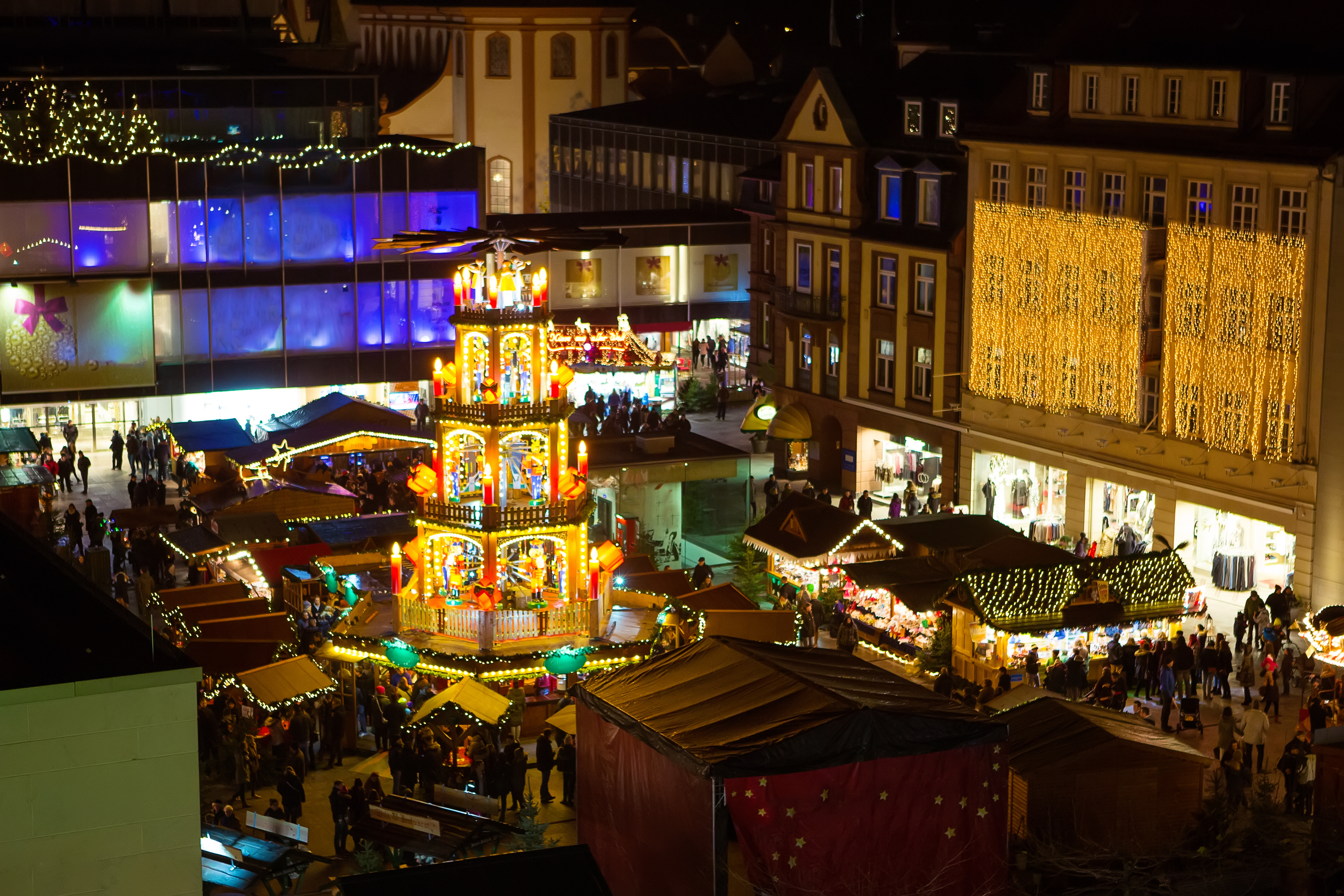 Mercado navideño nocturno, pirámide iluminada, puestos festivos y gente.