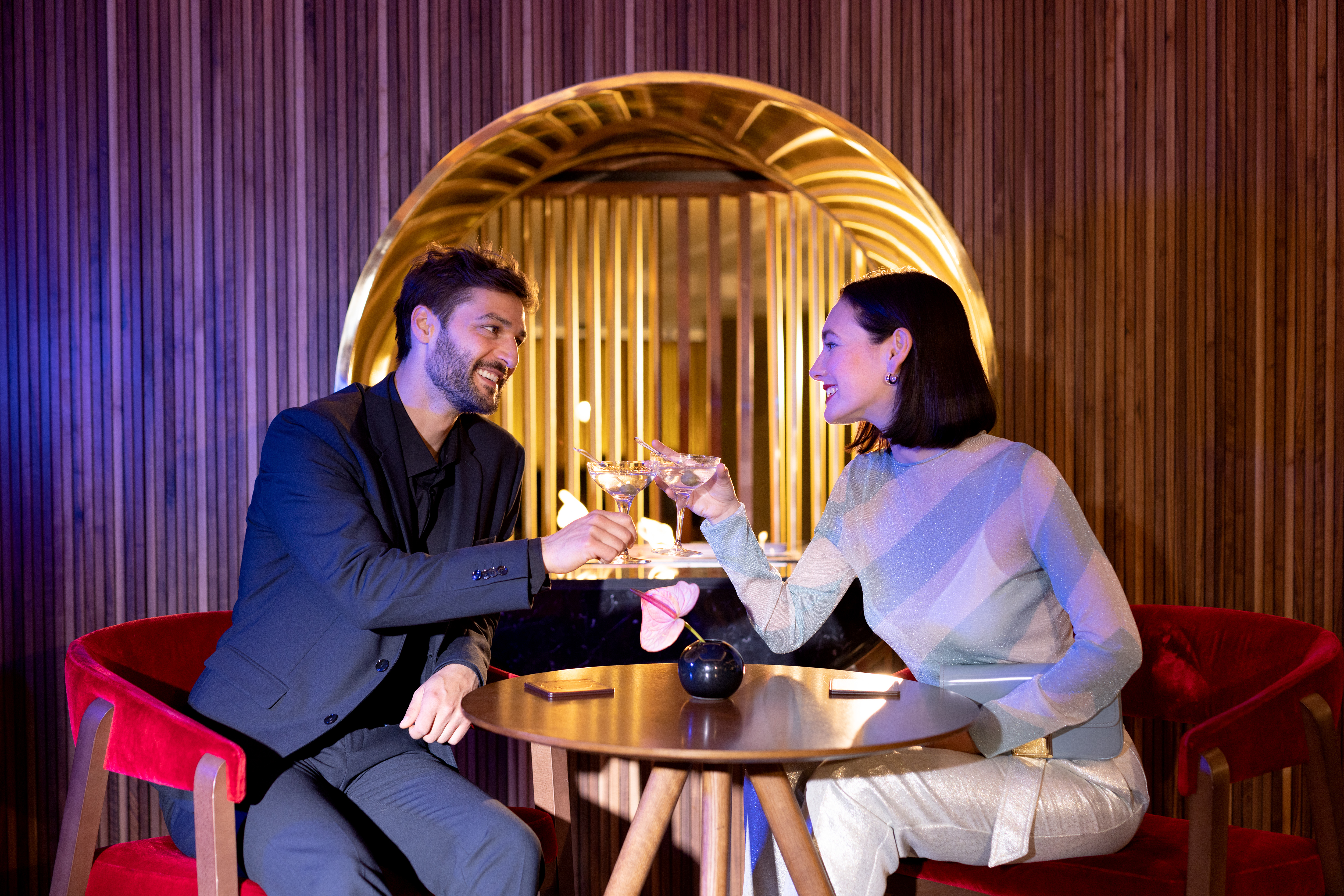 Smiling couple toasting cocktails in an elegant bar with a golden mirror.