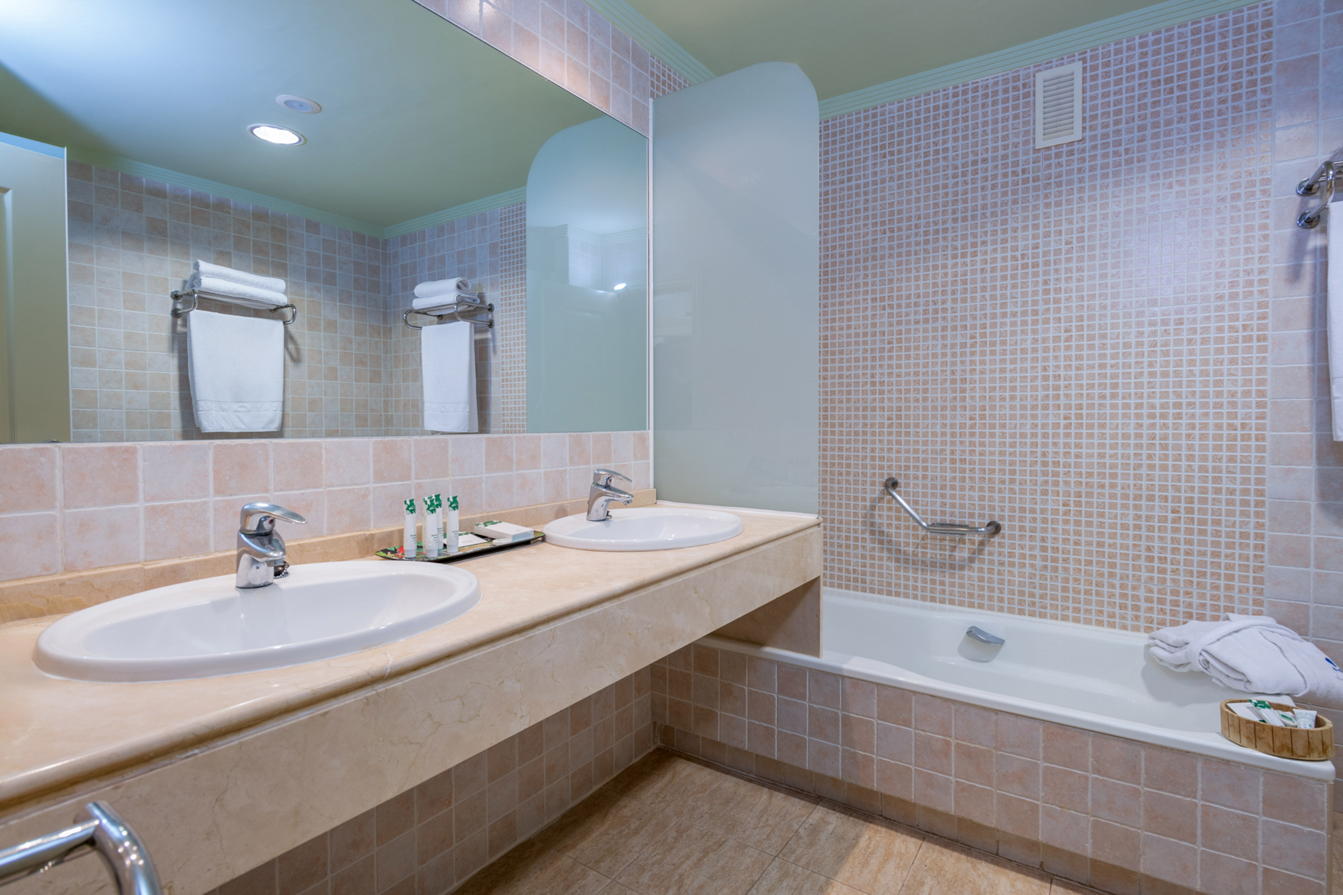 Clean hotel bathroom with double vanity, bathtub, mirror, and beige tiled walls.