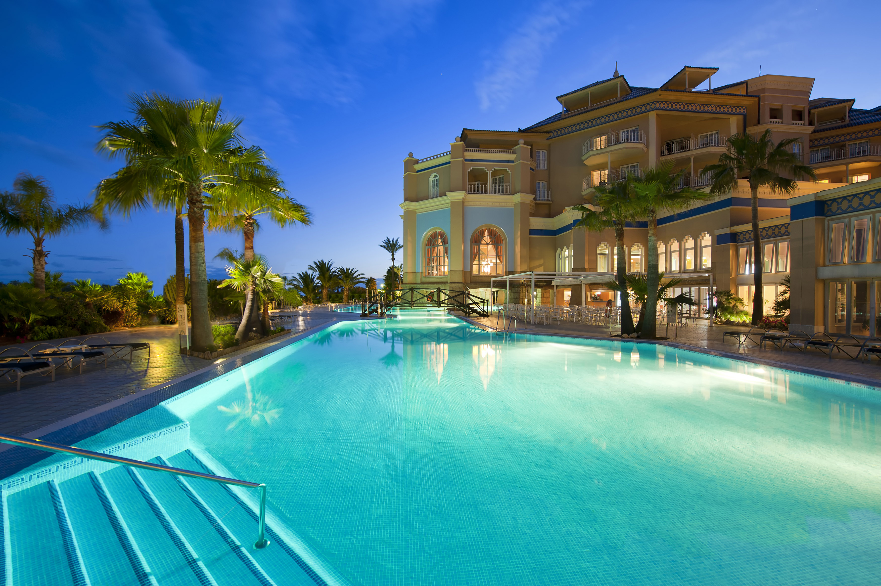 Illuminated luxury hotel pool, palm trees, and outdoor dining at dusk.