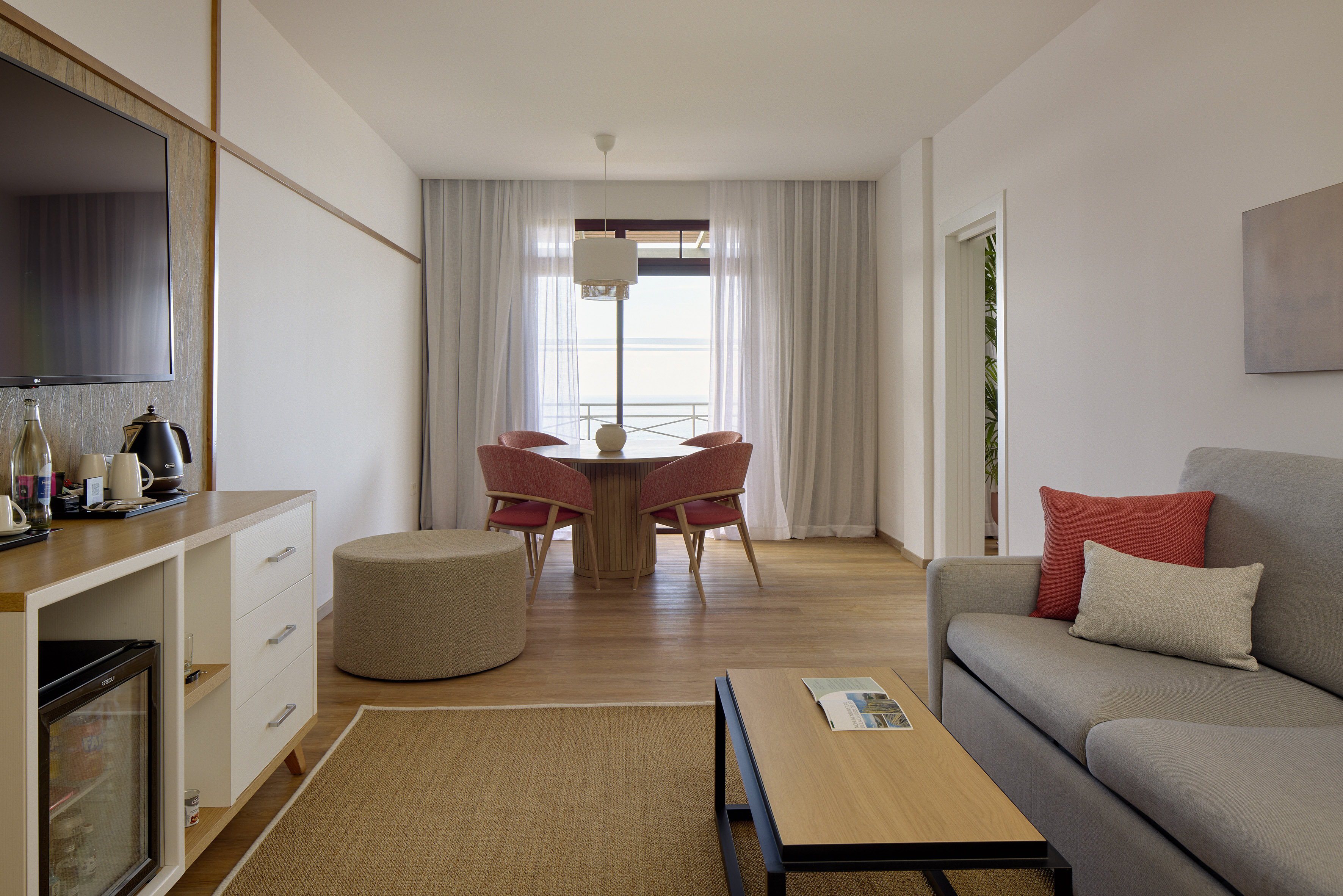 Modern hotel suite with dining table, grey sofa, and window ocean view.