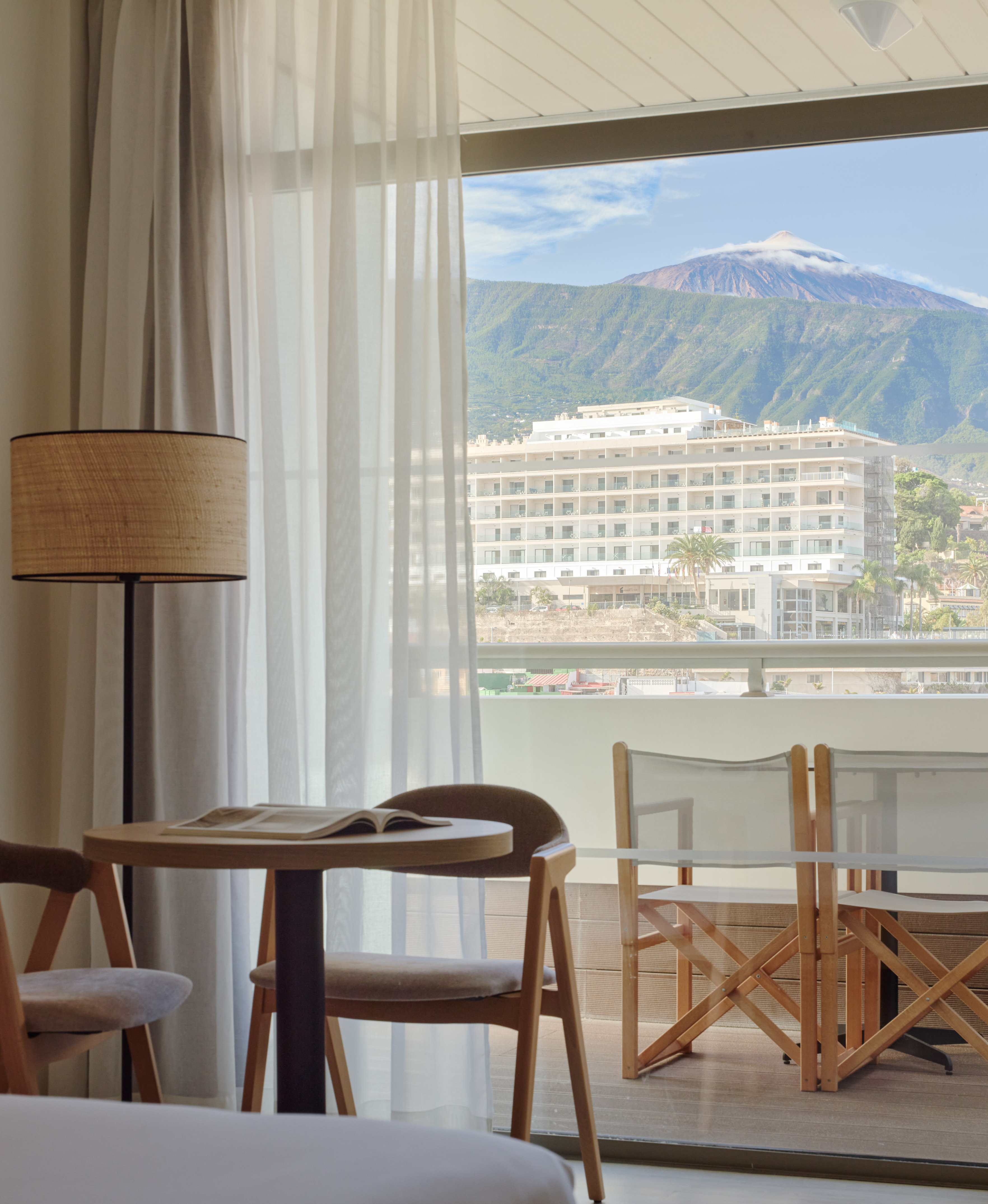 Hotel room with volcano mountain view from balcony. Resort building, table, chairs, lamp.
