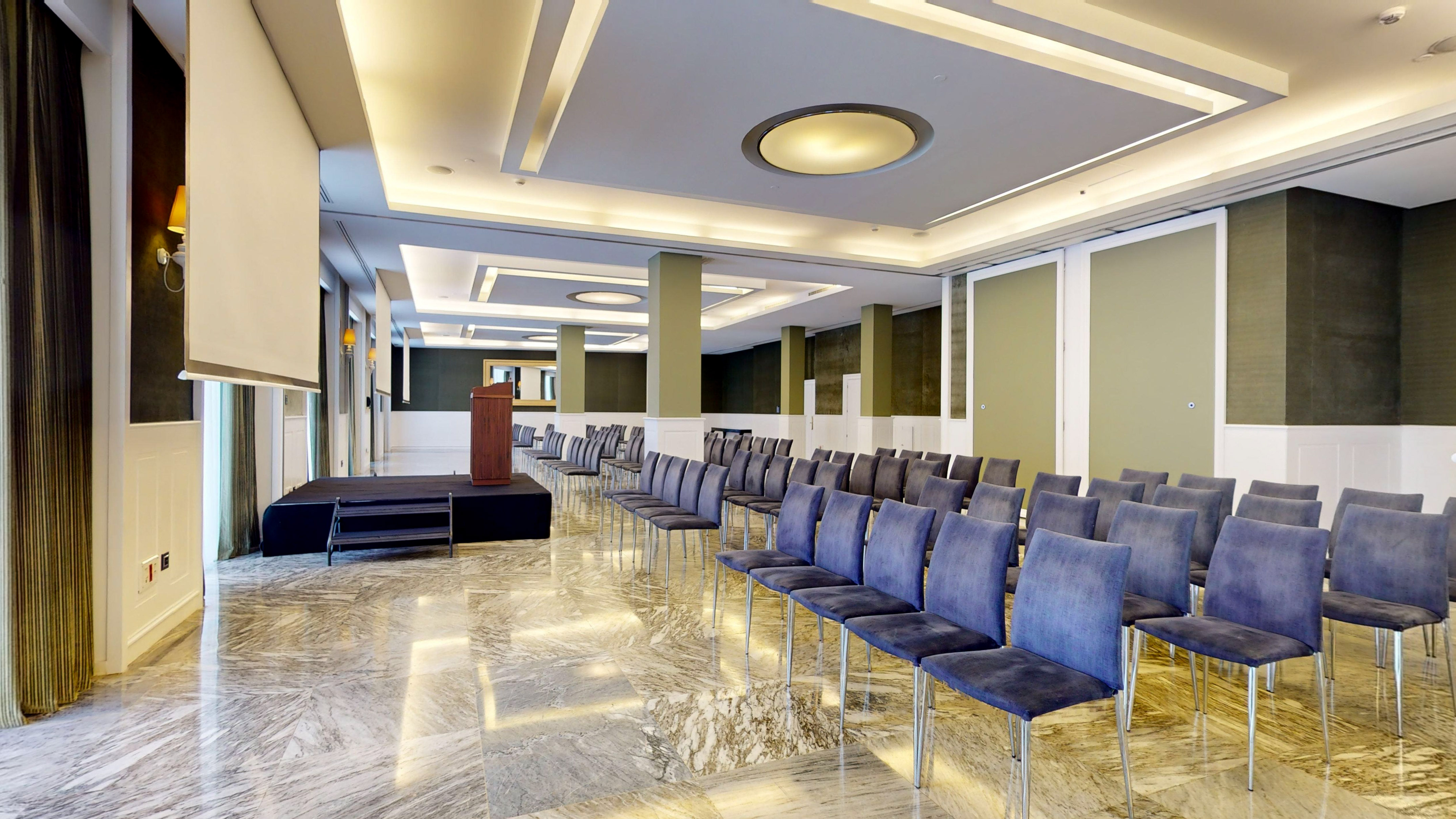 Spacious conference room with blue chairs, stage, podium, and projector screen.