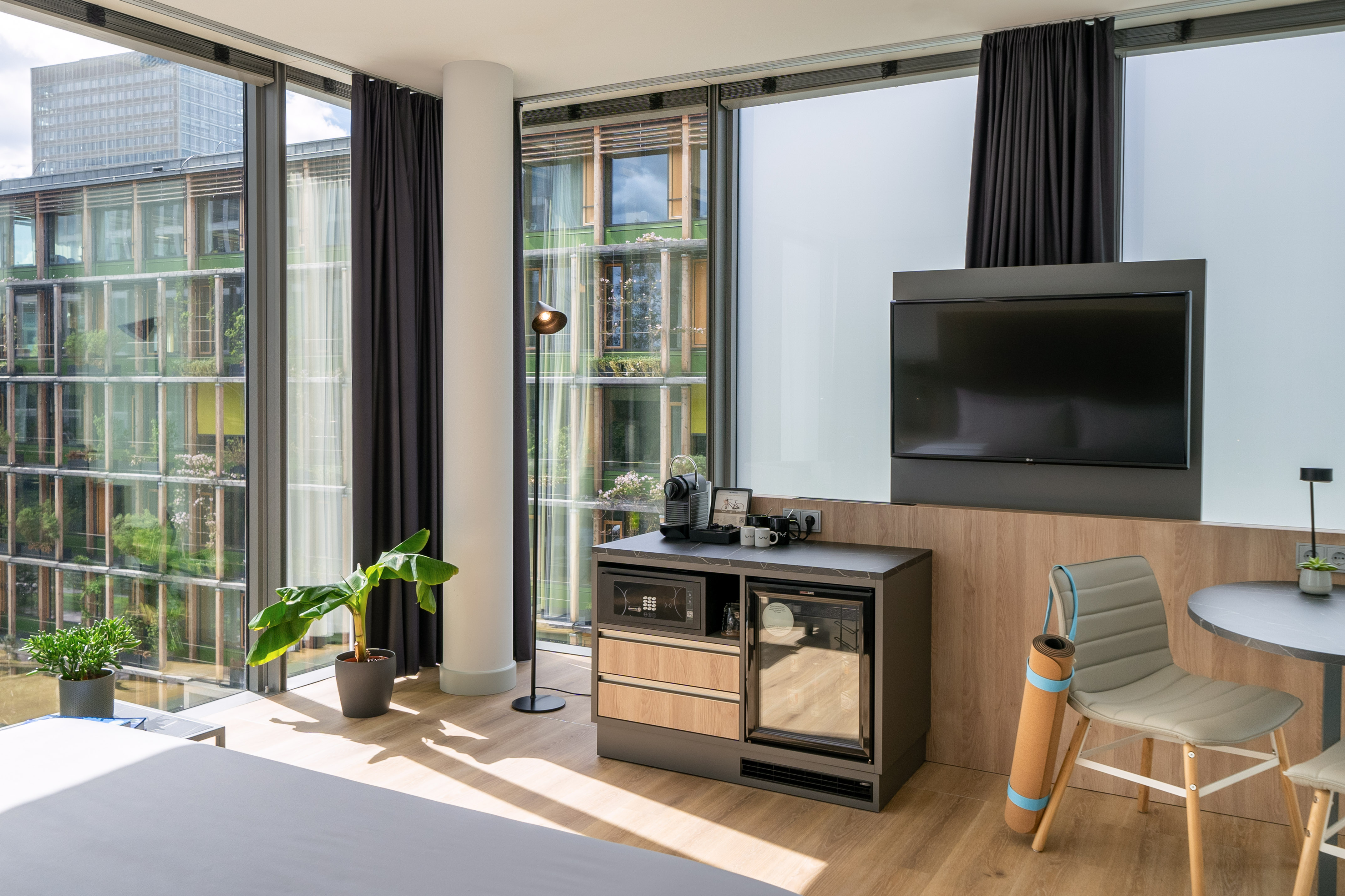 Modern hotel room with city view, TV, Nespresso, mini-fridge, and yoga mat.