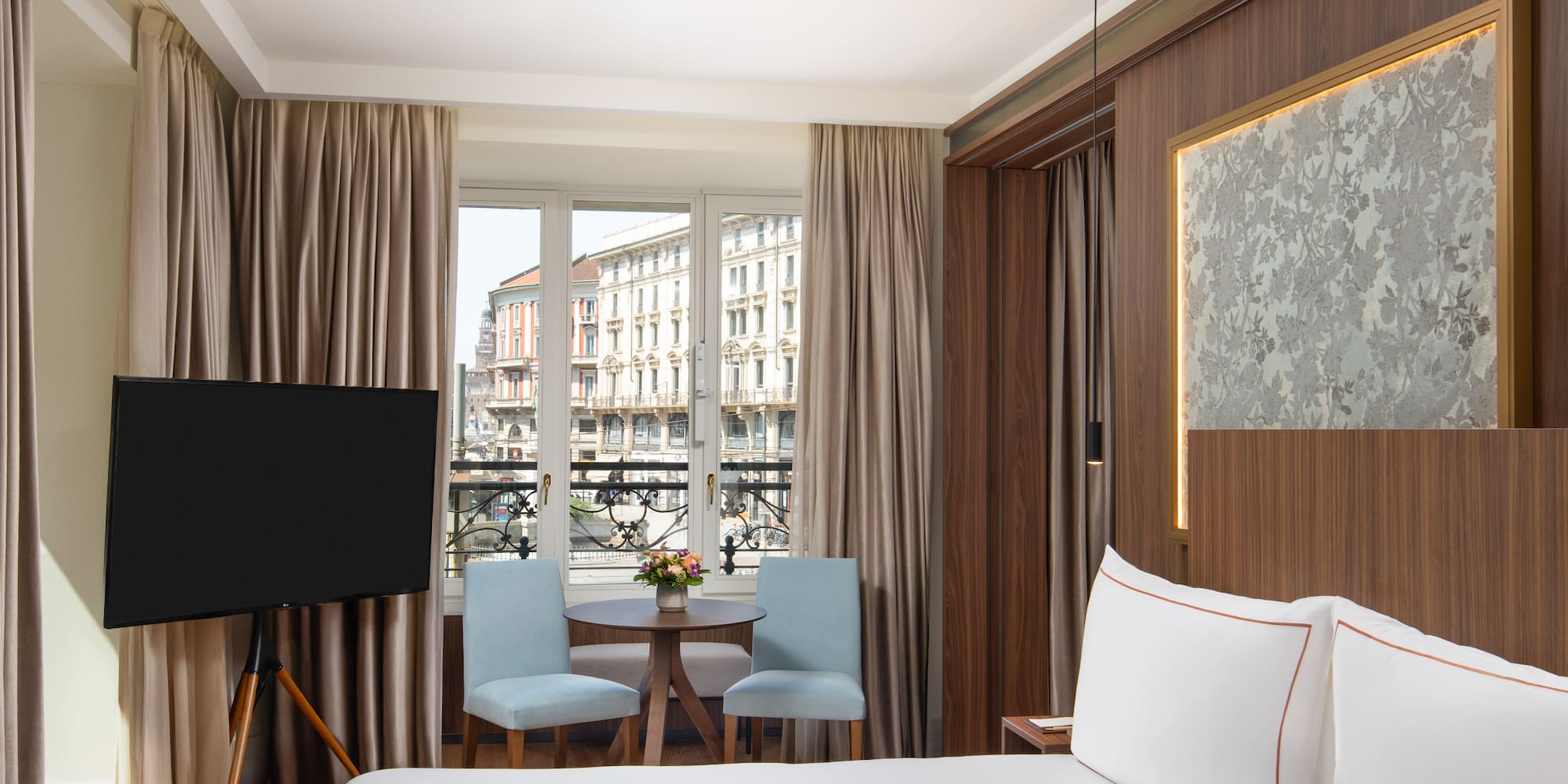 Hotel bedroom with city view, white bed, blue chairs, and flat-screen TV.