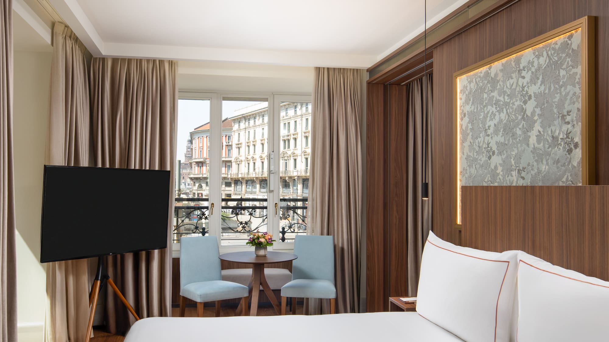 Hotel bedroom with city view, white bed, blue chairs, and flat-screen TV.