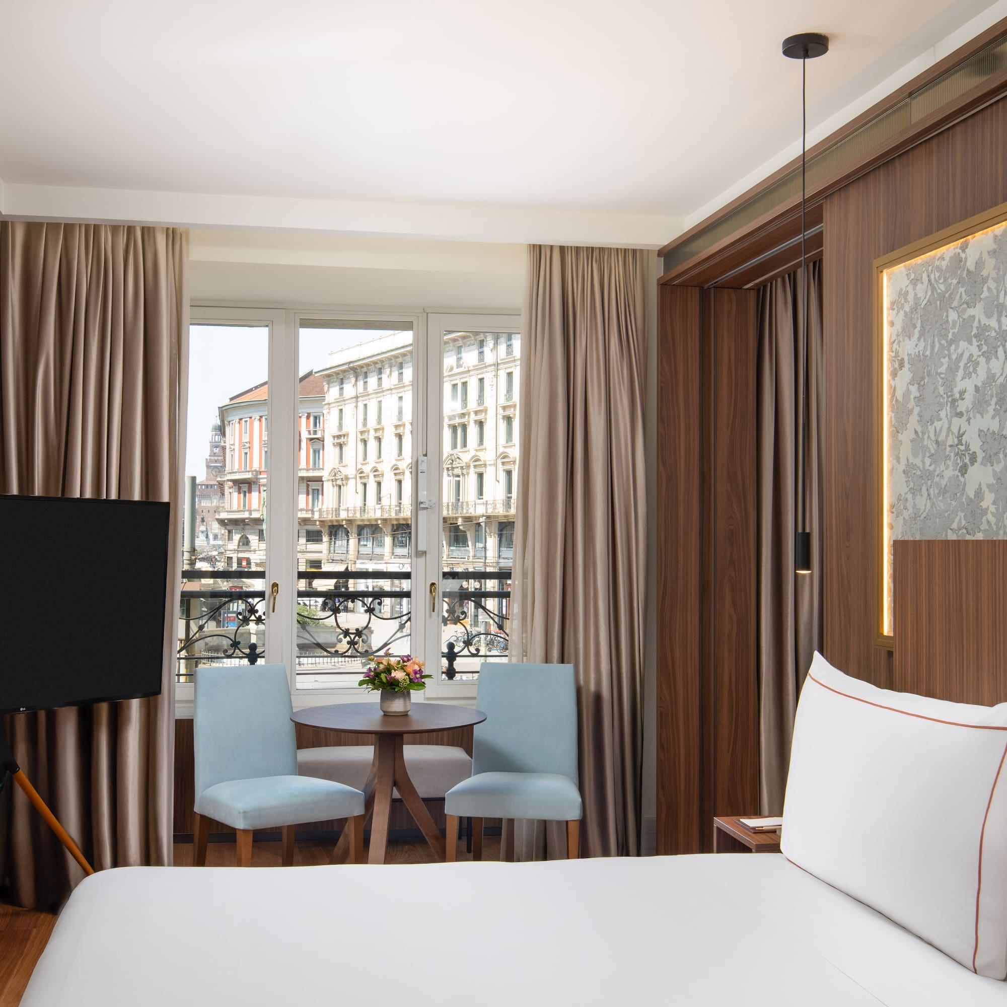 Hotel bedroom with city view, white bed, blue chairs, and flat-screen TV.