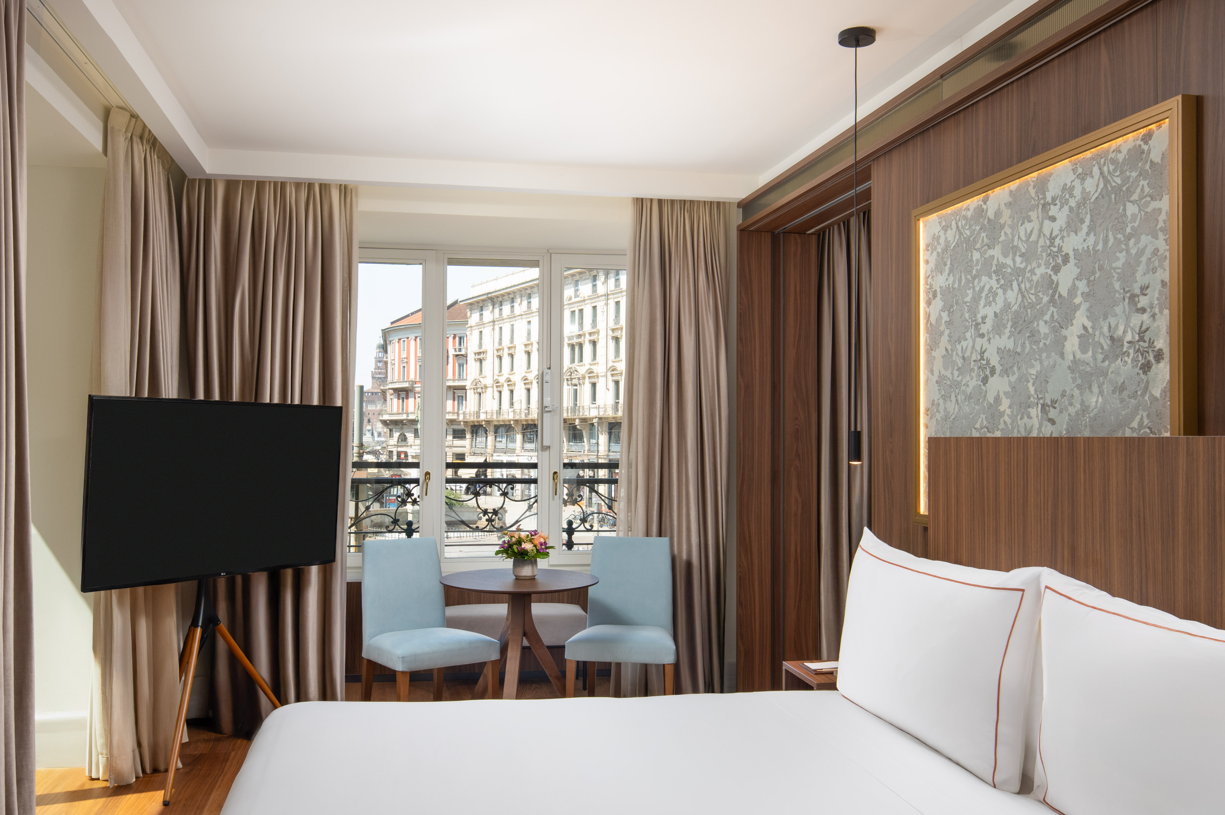 Hotel bedroom with city view, white bed, blue chairs, and flat-screen TV.