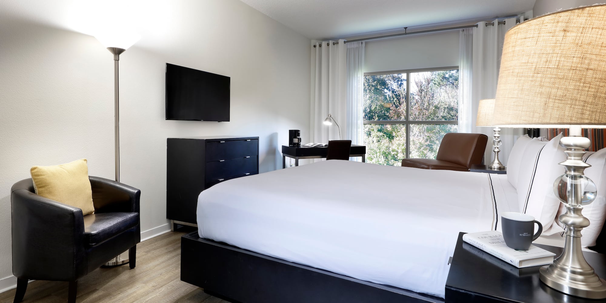 Modern hotel bedroom with king bed, workstation, TV, and large window.