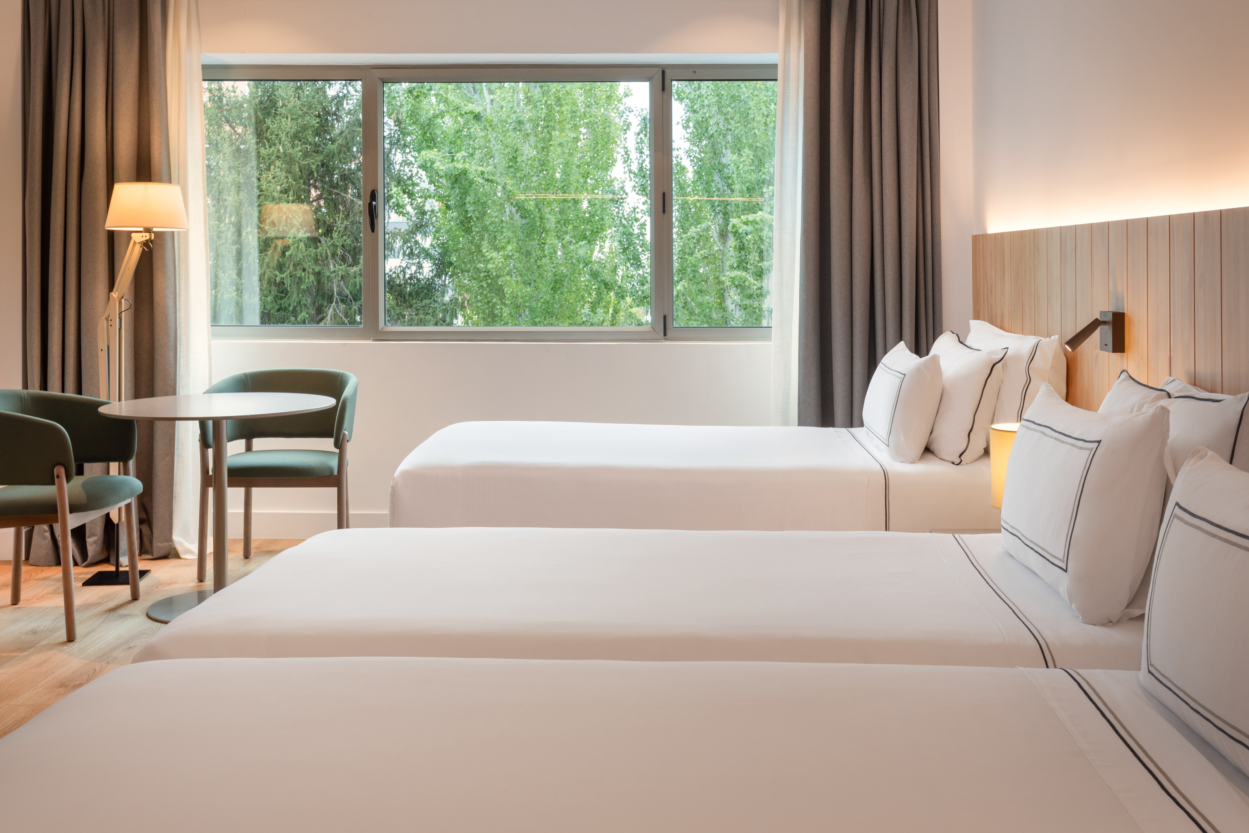 Hotel room: three twin beds, green chairs, window with lush tree view.