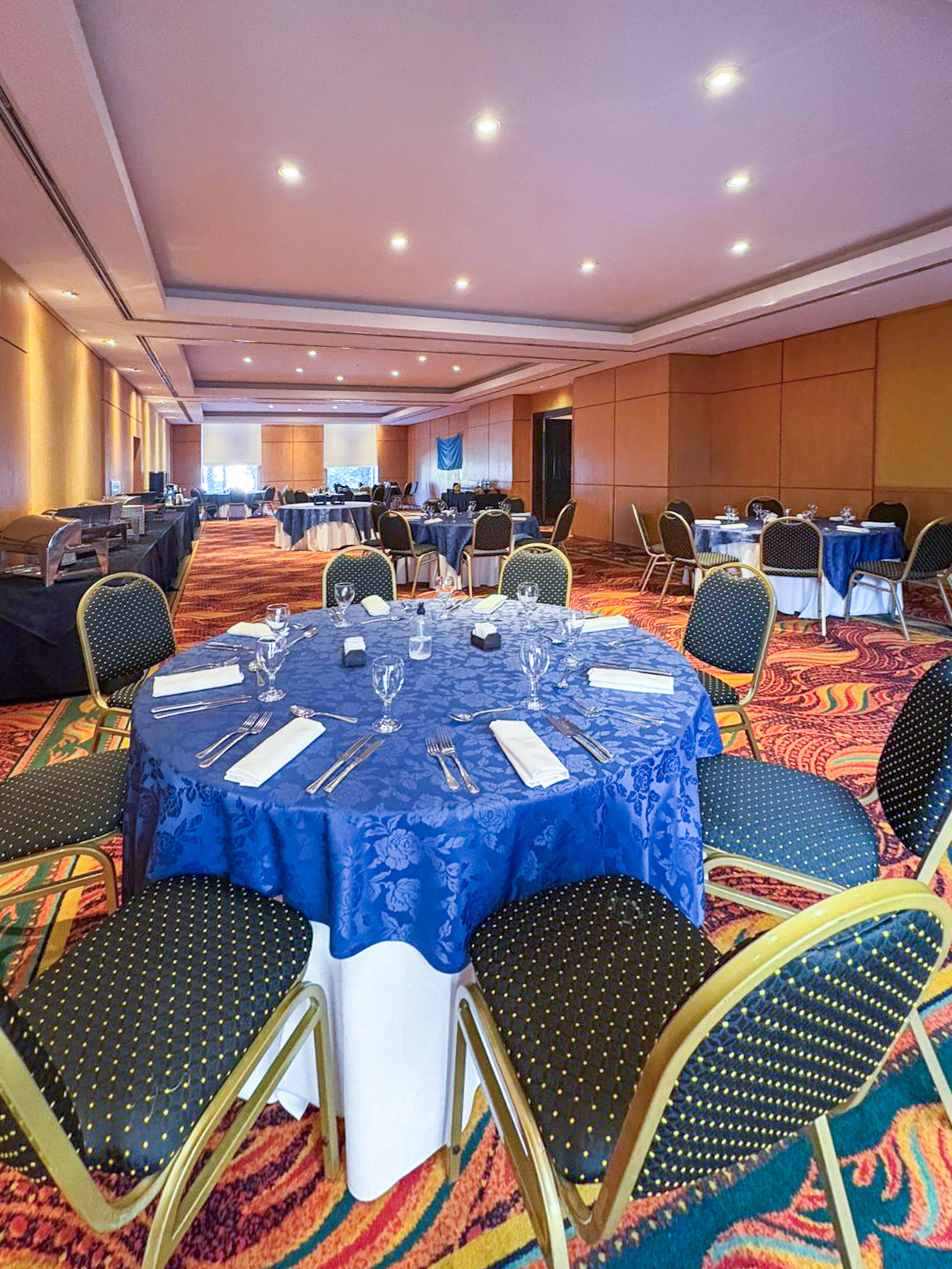 Hotel banquet hall with blue-clothed round tables, chairs, and buffet stations.