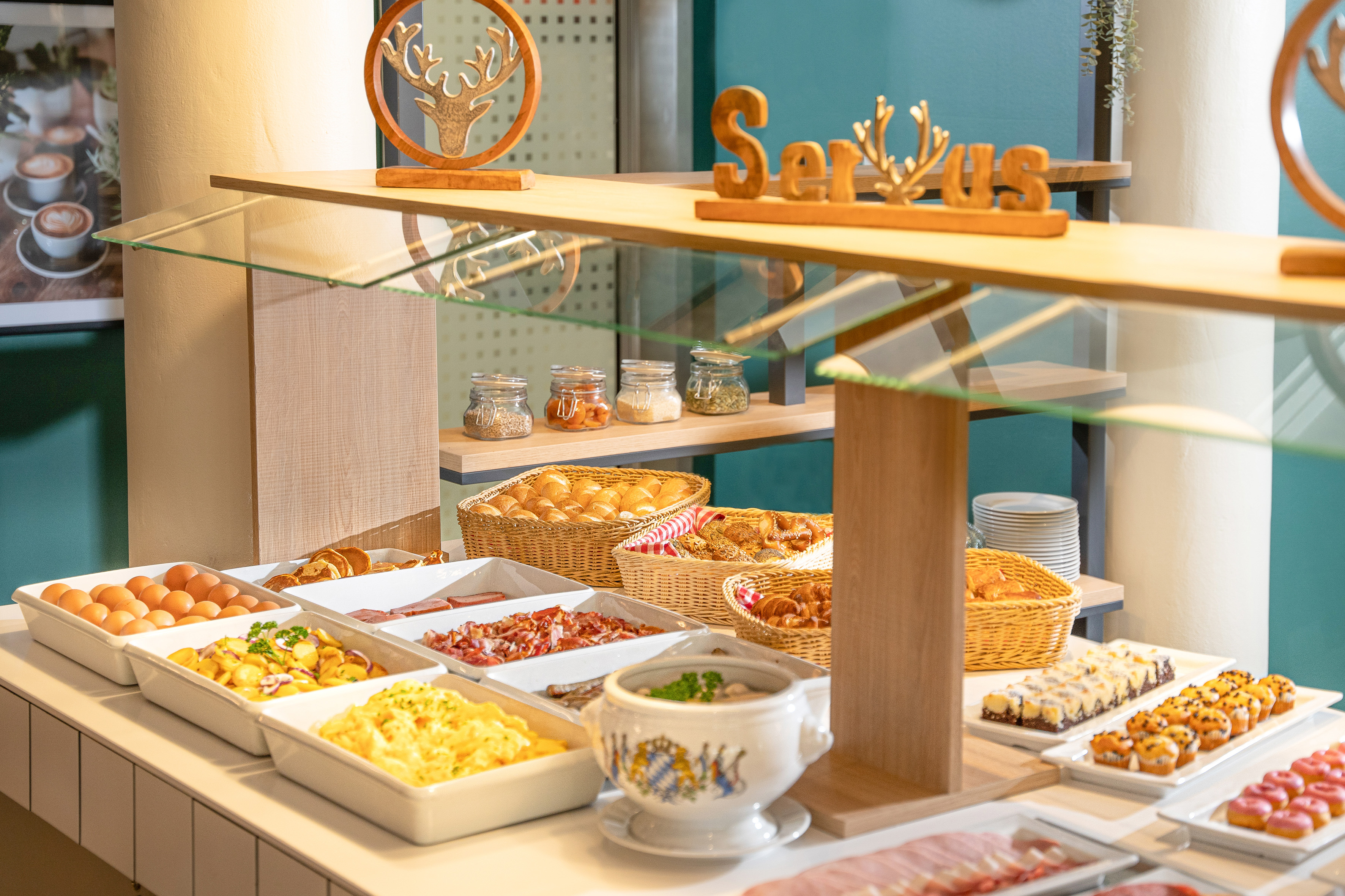 Breakfast buffet with eggs, bacon, pastries, bread, and "Servus" sign.