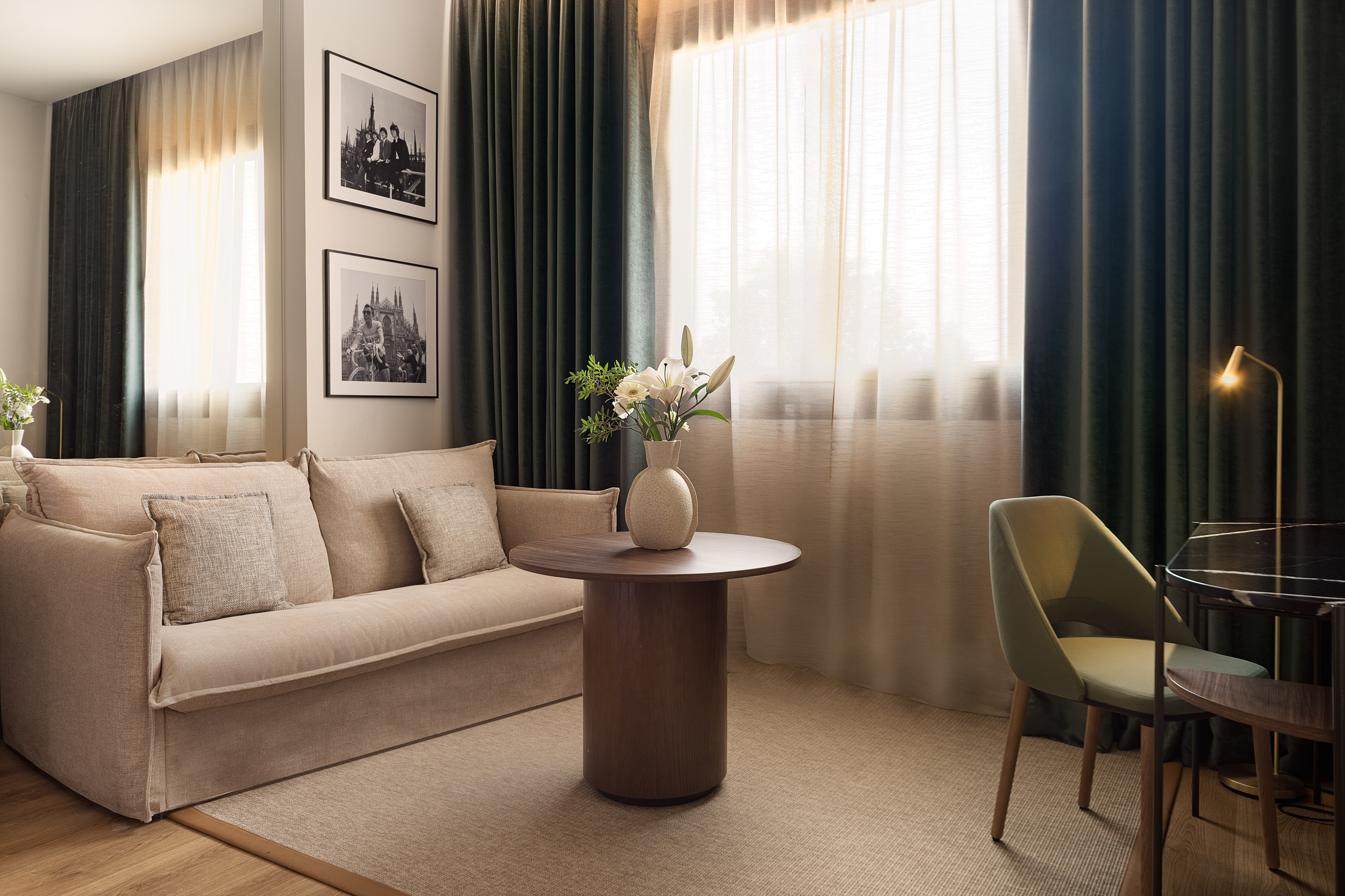 Modern hotel room with beige sofa, wood table, green chair, and dark curtains.