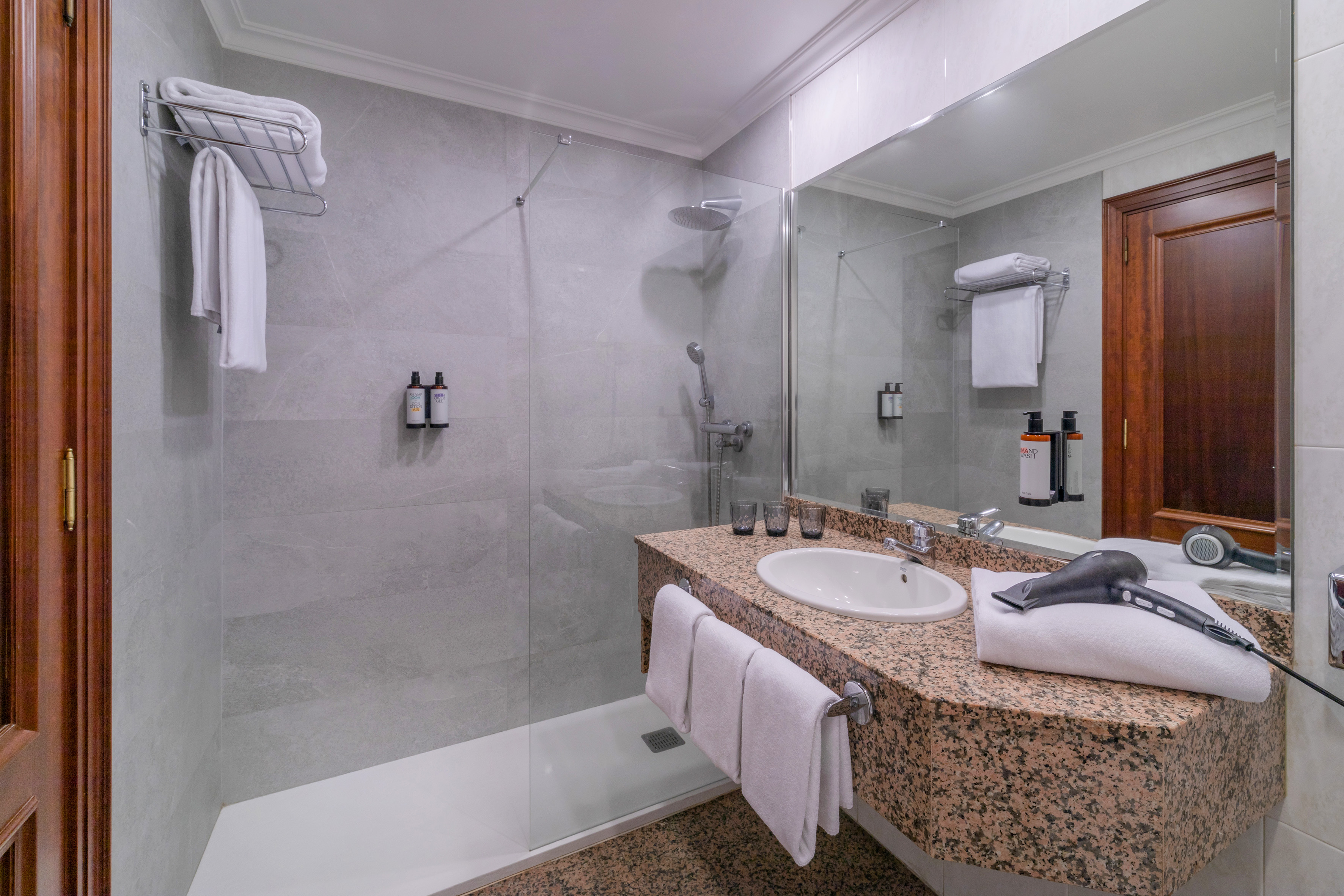 Bright hotel bathroom with granite vanity, sink, walk-in shower, and hairdryer.