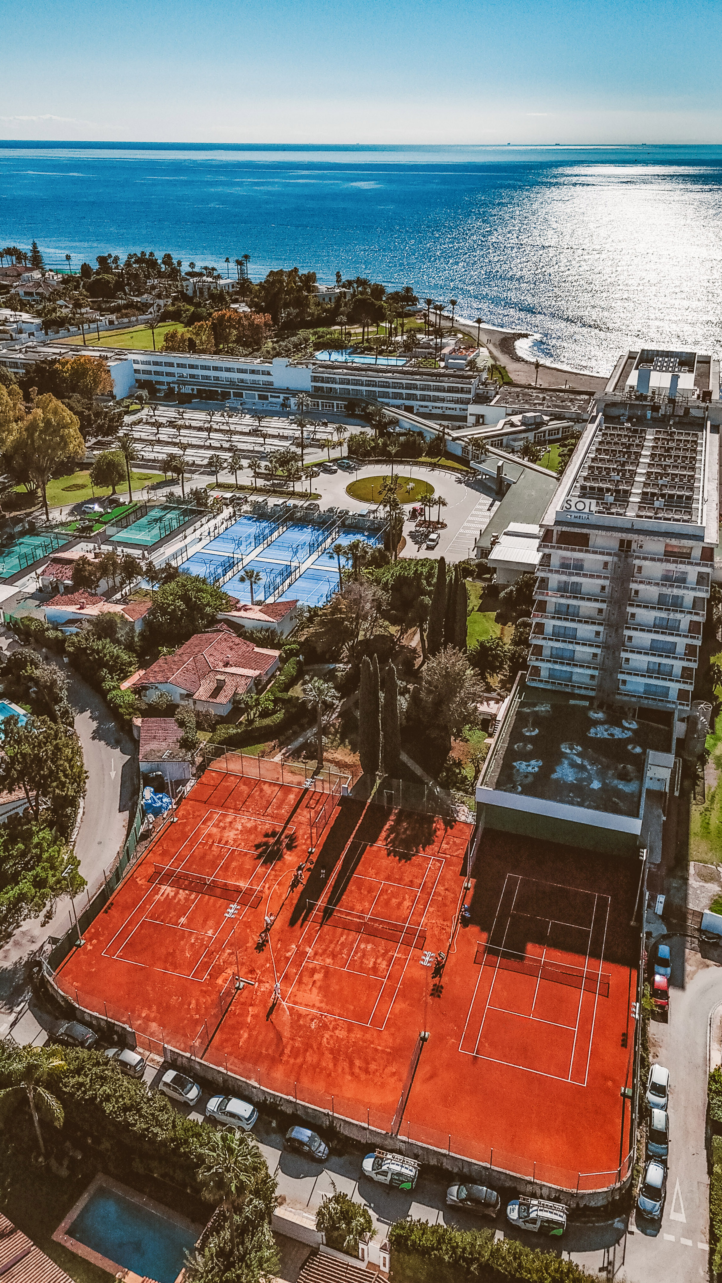 Aerial view of coastal resort with red clay tennis, blue padel courts, ocean.