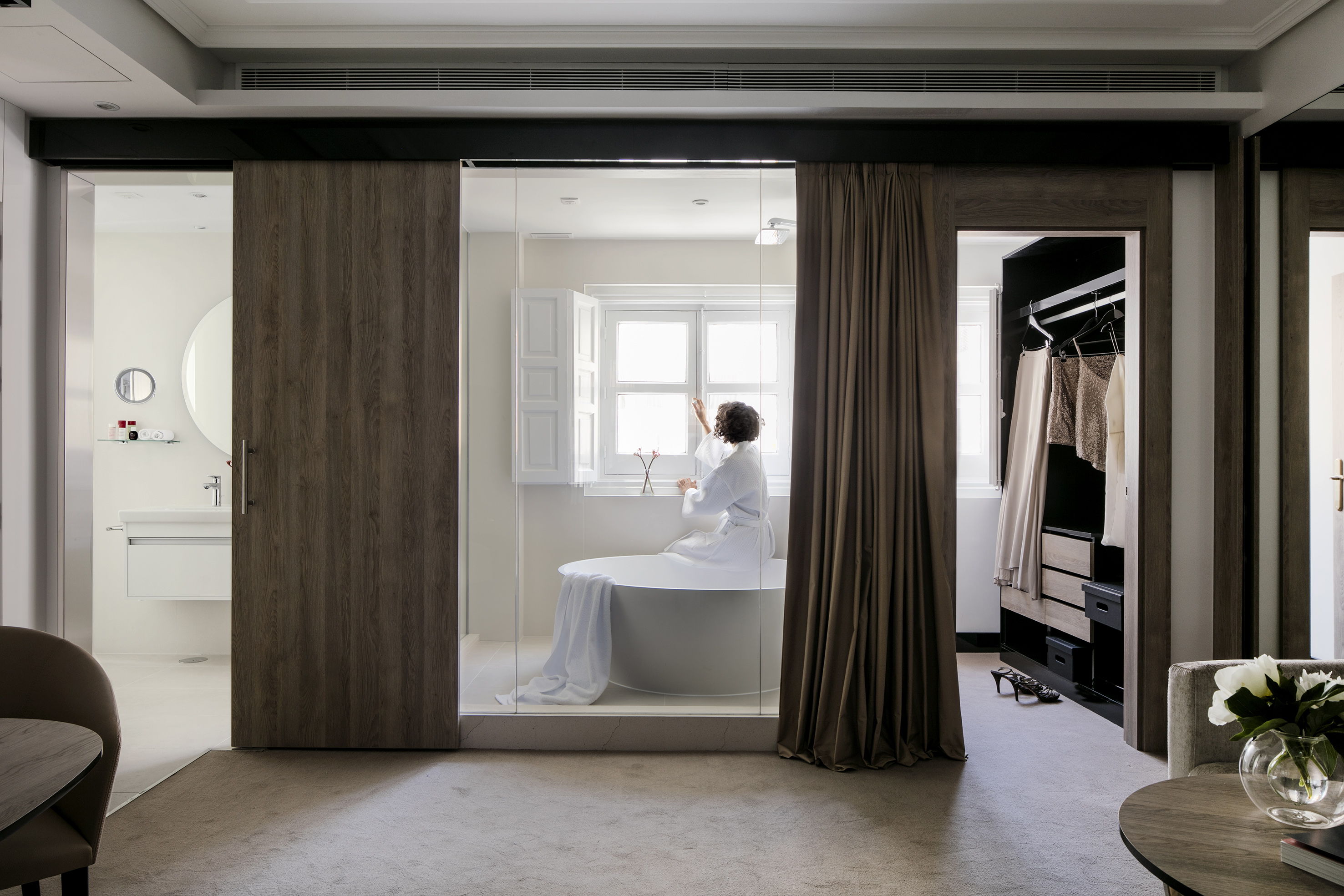 Woman in bathrobe by modern freestanding tub in luxury hotel suite.