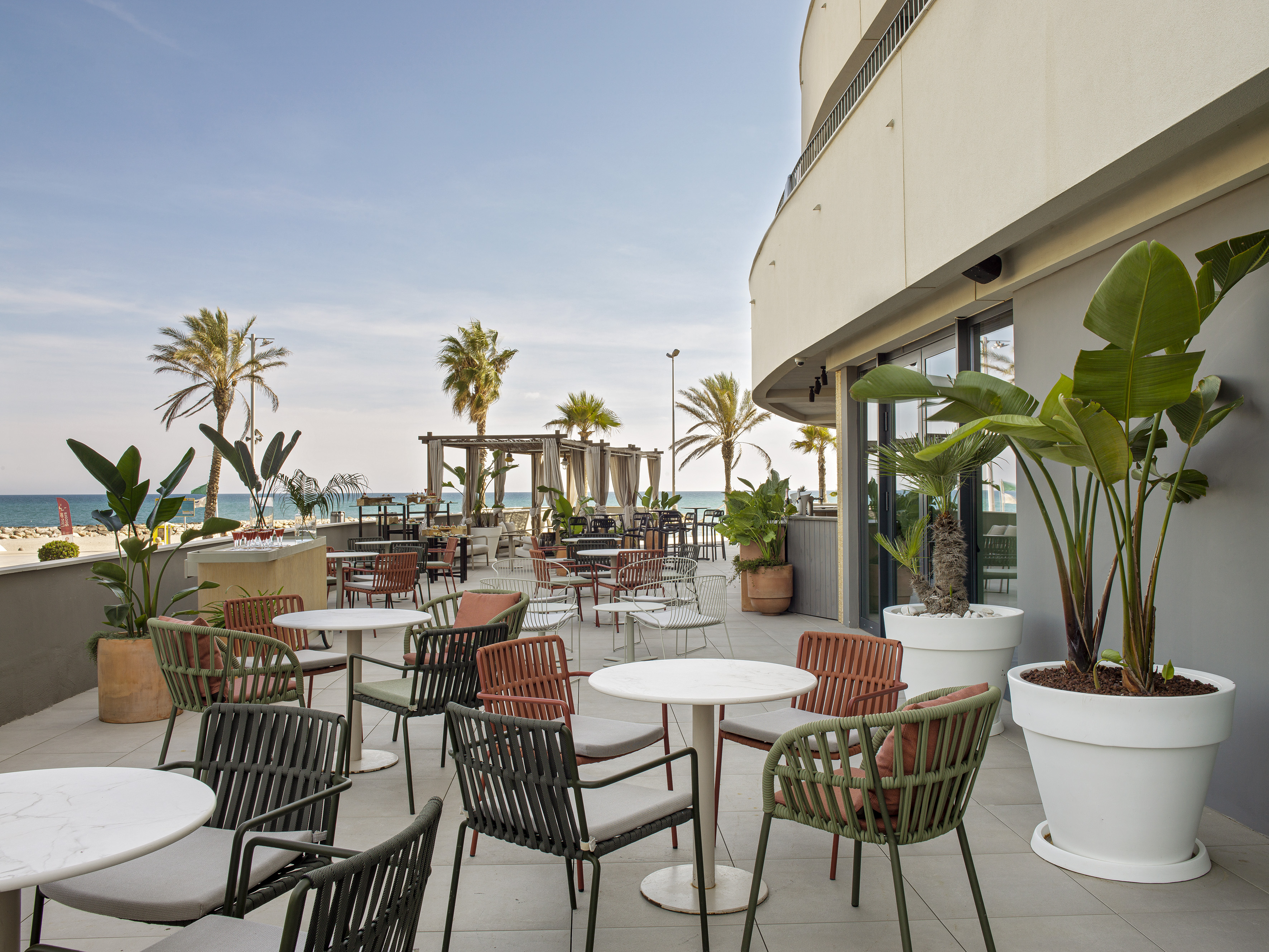 Beachfront hotel patio with modern dining tables, palm trees, and ocean view.