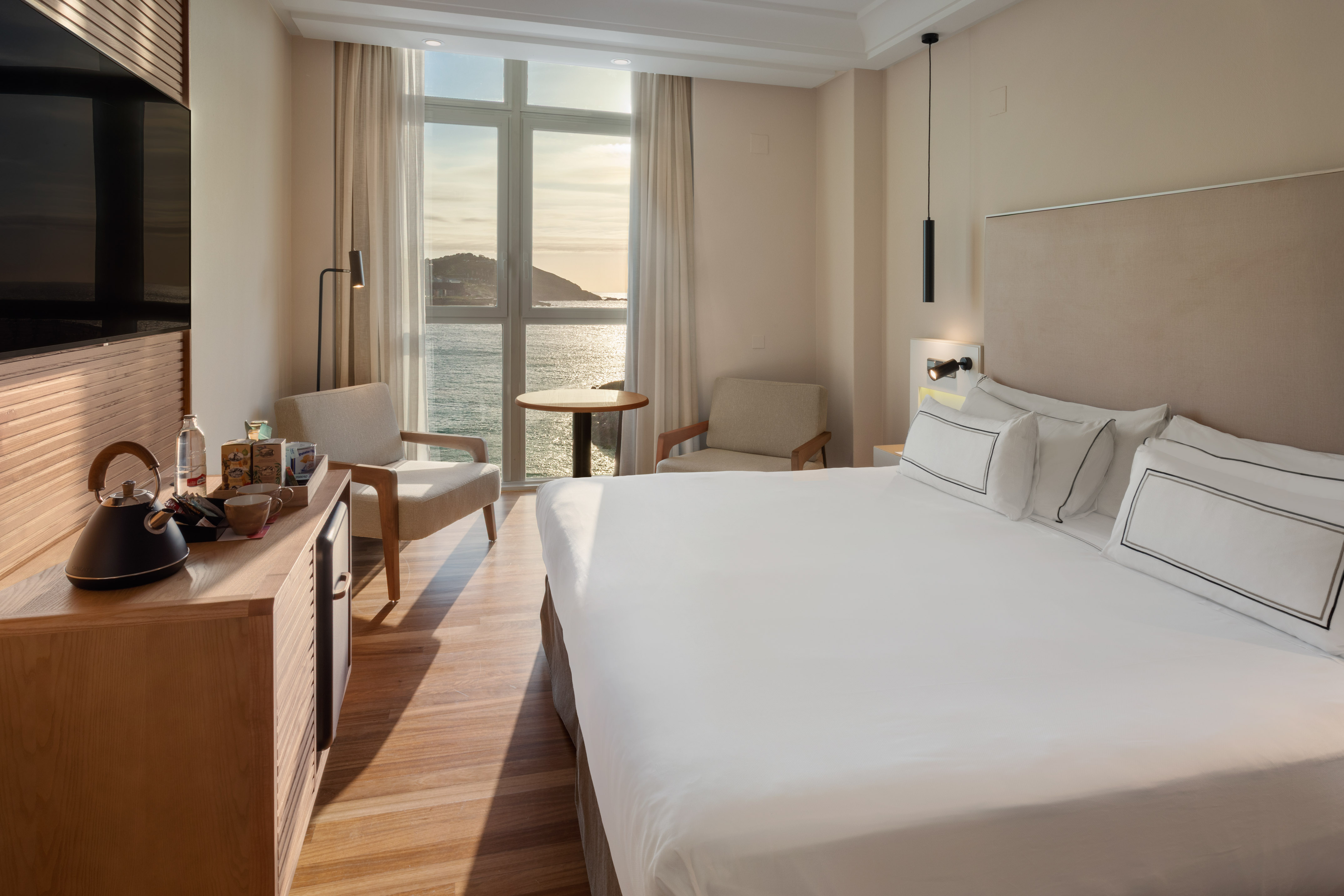 Modern hotel room with ocean view, large bed, and cozy seating by the window.