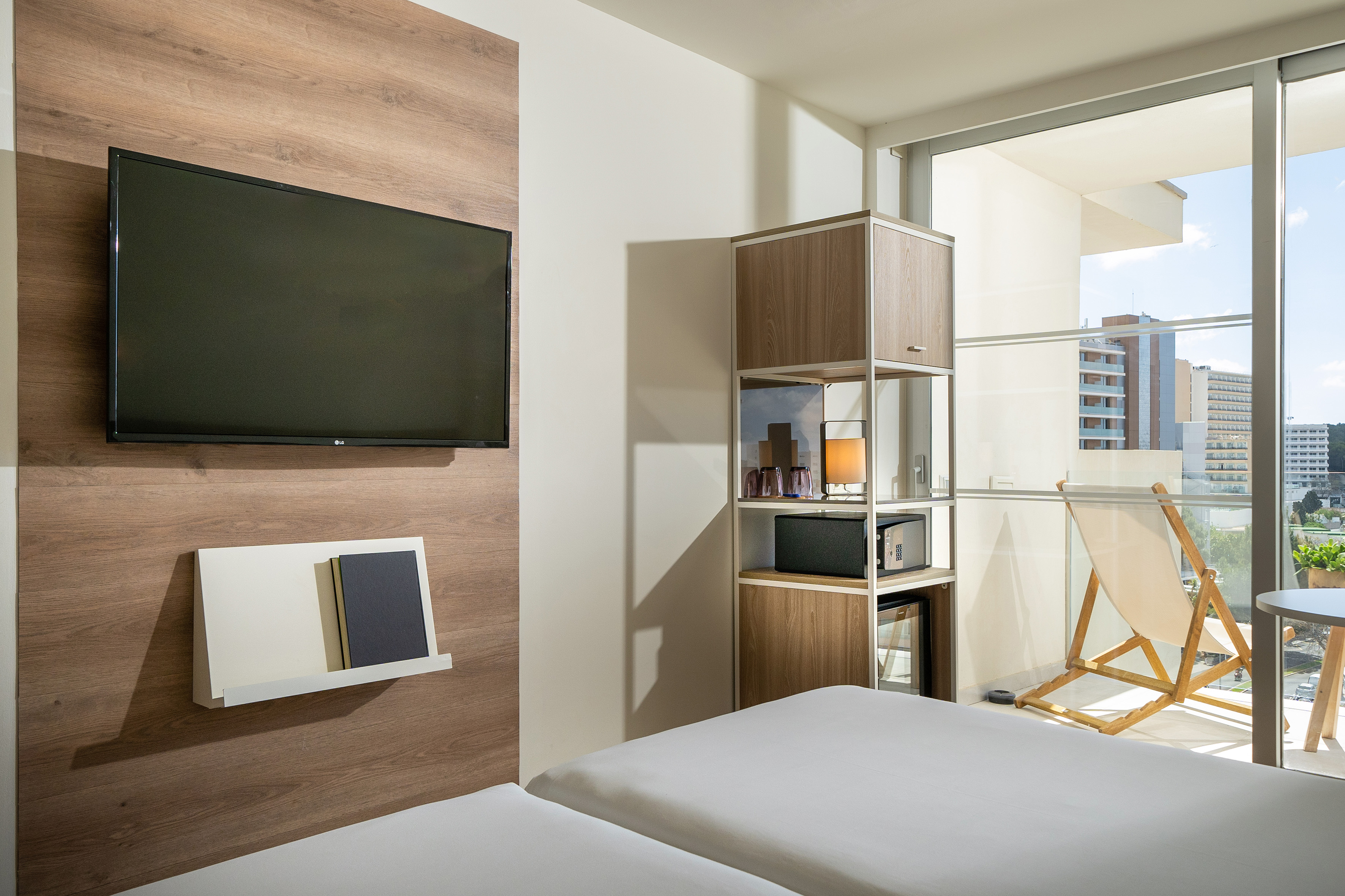 Modern hotel bedroom with twin beds, TV, mini-fridge, and balcony city view.