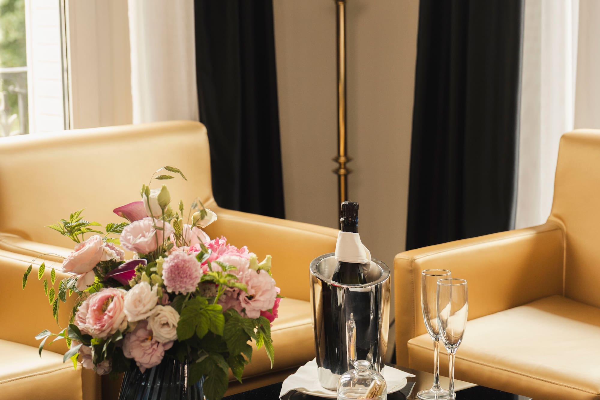 Elegant hotel room with champagne, flowers, macarons, and tan armchairs.