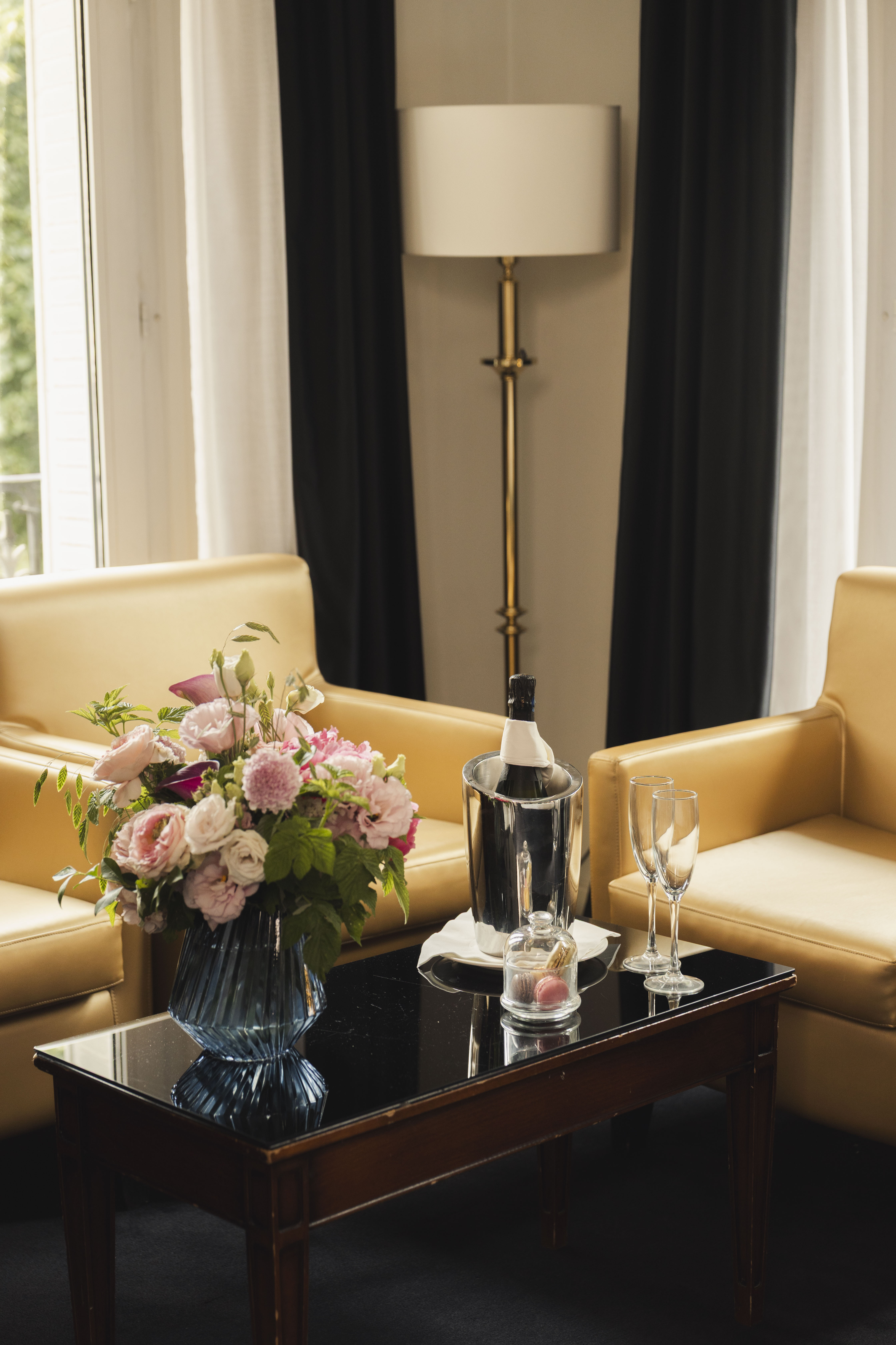 Elegant hotel room with champagne, flowers, macarons, and tan armchairs.